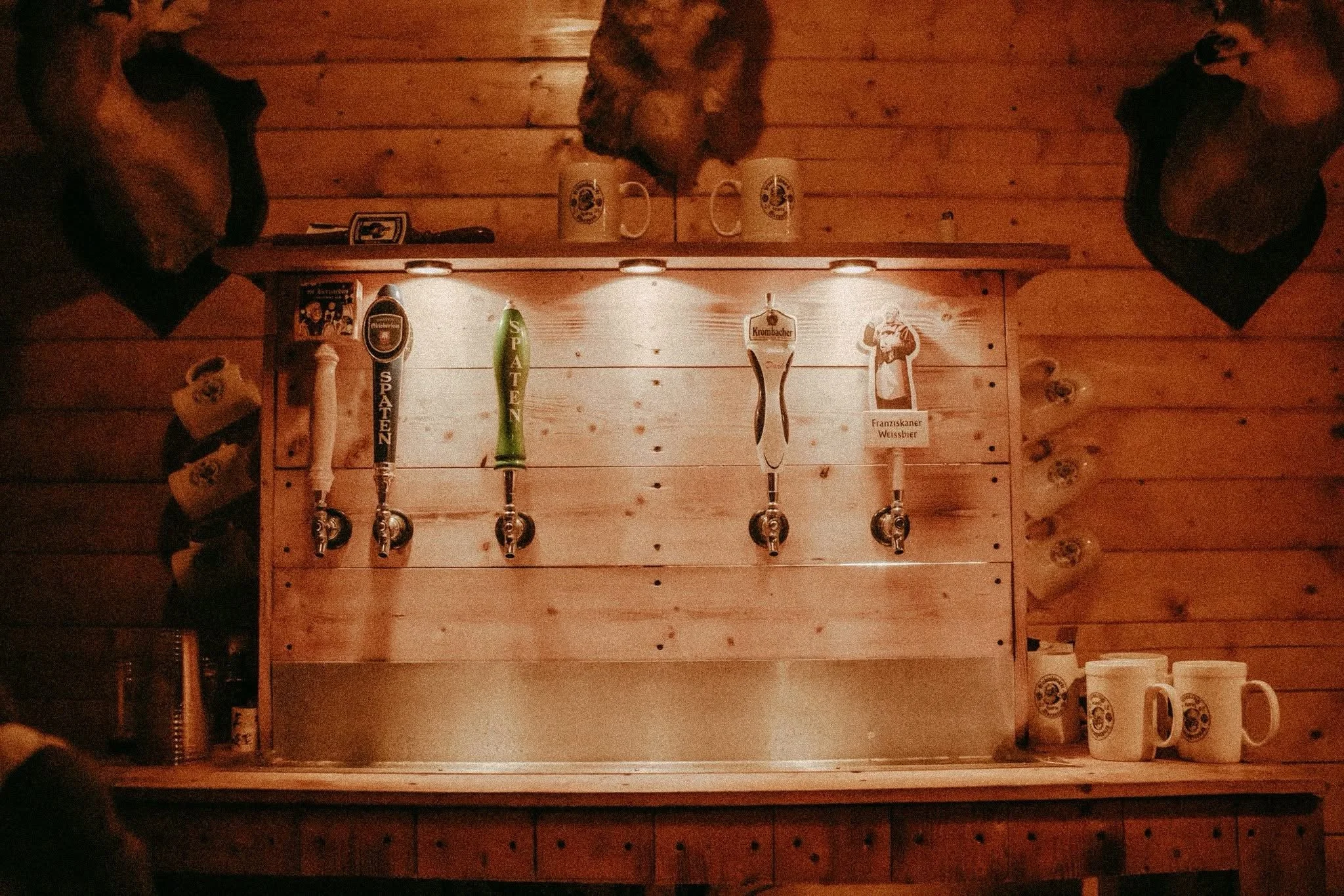 A wooden bar area with five beer taps, three of which are labeled with different beer brands, and  two mugs of beer with foam on top. The bar is illuminated by ceiling lights, and there are additional mugs on the right side of the bar. The background features a wooden wall with some objects and decorations.