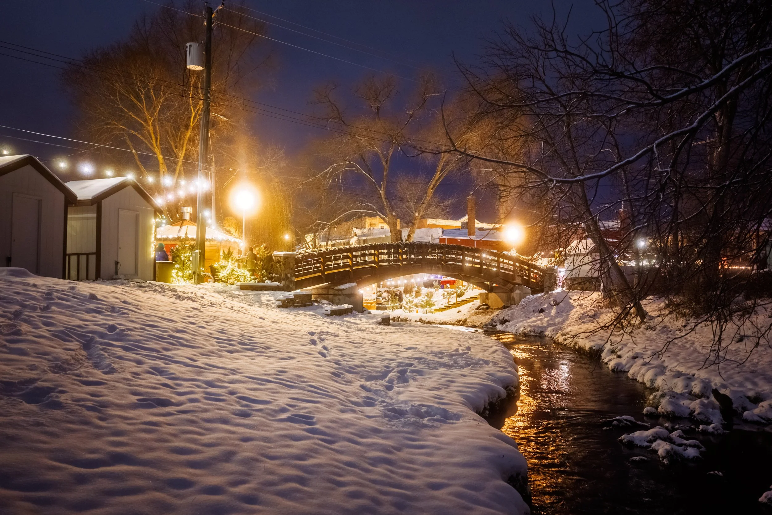 A snowy scene at night with snow-covered ground, a small river, wooden bridge with holiday lights, and houses in the background. Many leafless trees are also visible.