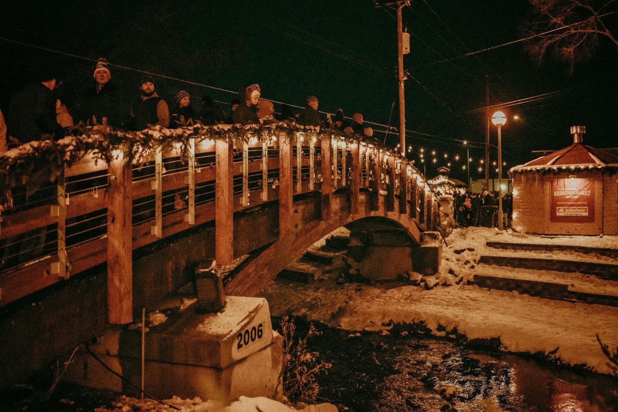 People standing on a snow-covered wooden bridge decorated with holiday lights at night, with a small snow-covered building and tall poles with lights in the background.