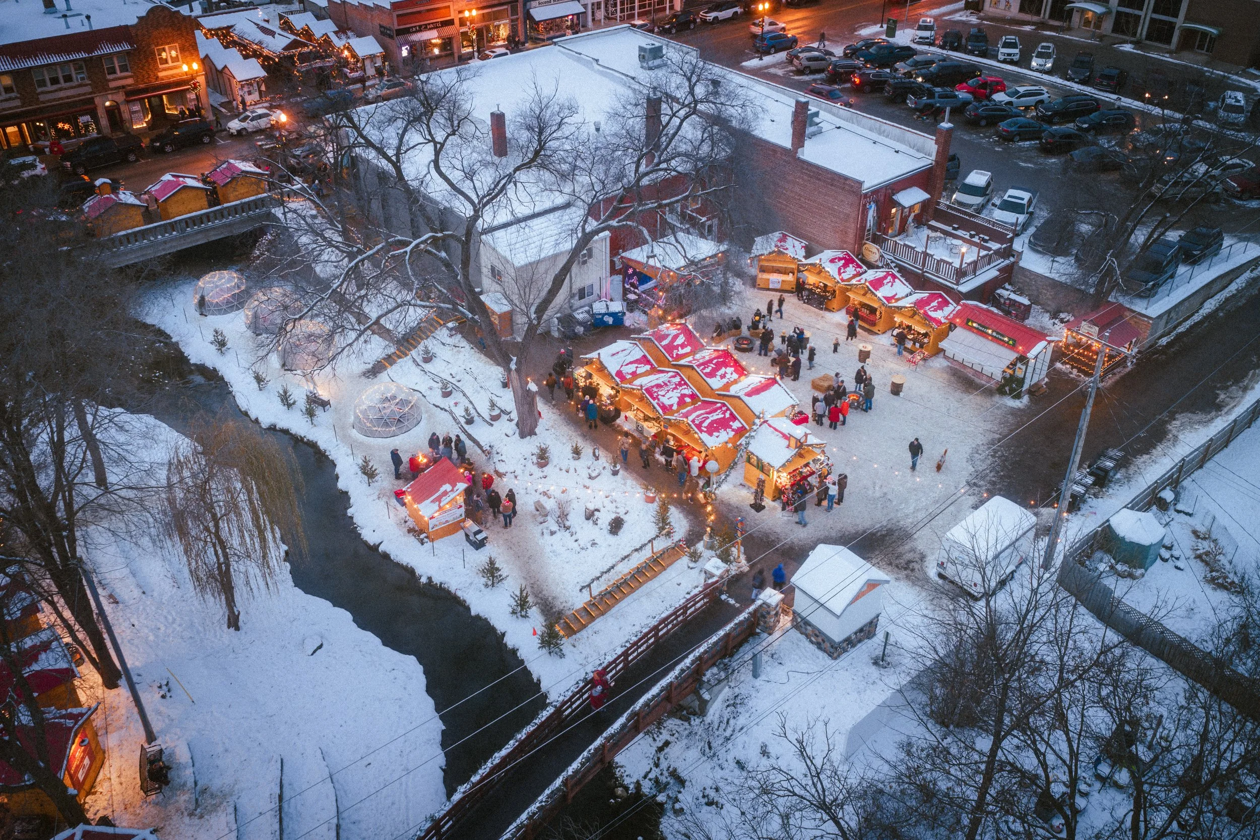 An aerial view of a small town winter market with various vendor stalls decorated with red and white striped awnings, people walking around, snow-covered rooftops, leafless trees, a narrow river, and parked cars along the street at dusk.