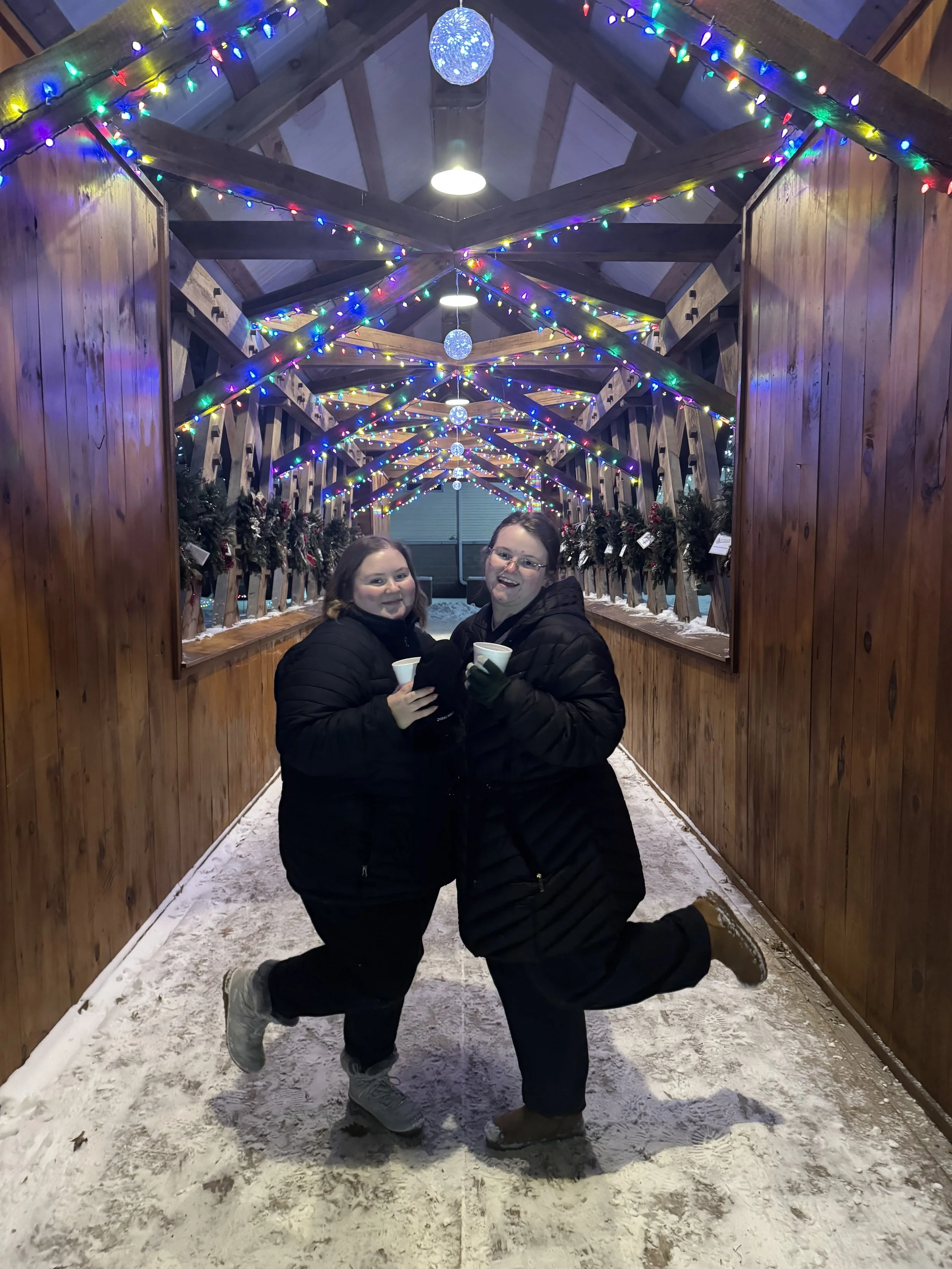 Two women wearing black jackets and gloves in a covered outdoor area decorated with colorful string lights and Christmas wreaths. They are smiling, with one woman kneeling and the other standing, holding cups, on snowy ground.