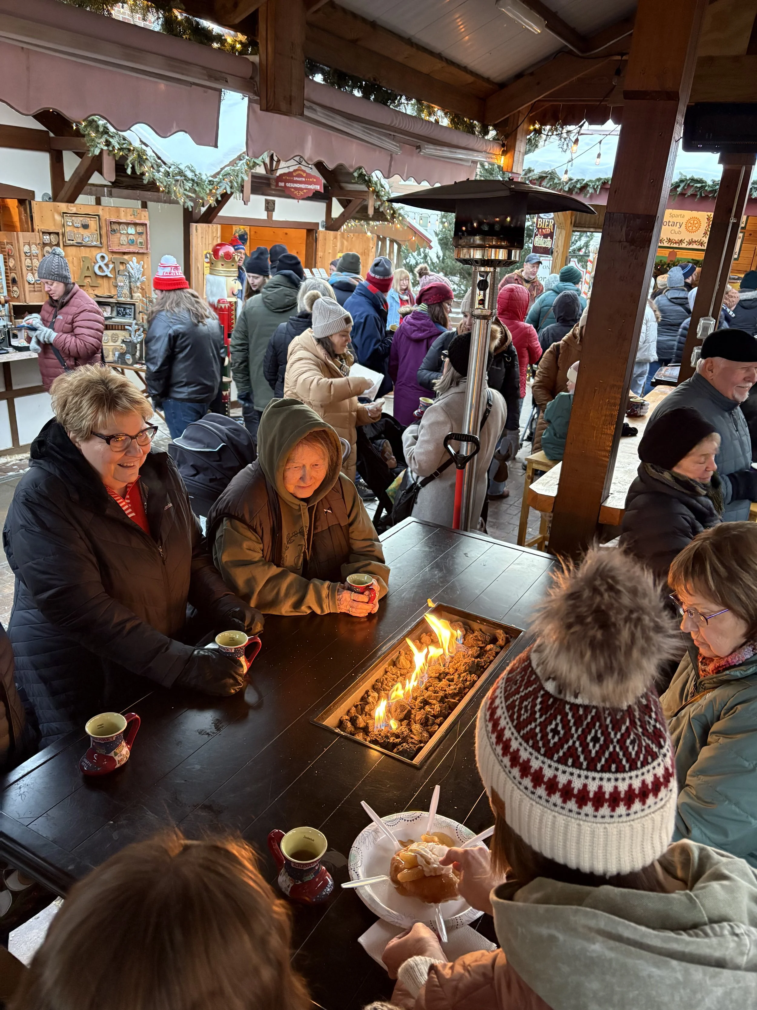 People gather around a long table at an outdoor winter market, sitting near a fire pit with flames. Many are wearing winter hats and jackets, and some are holding cups of hot drinks. The market has wooden stalls decorated with holiday ornaments and greenery, with a crowd of shoppers in the background.