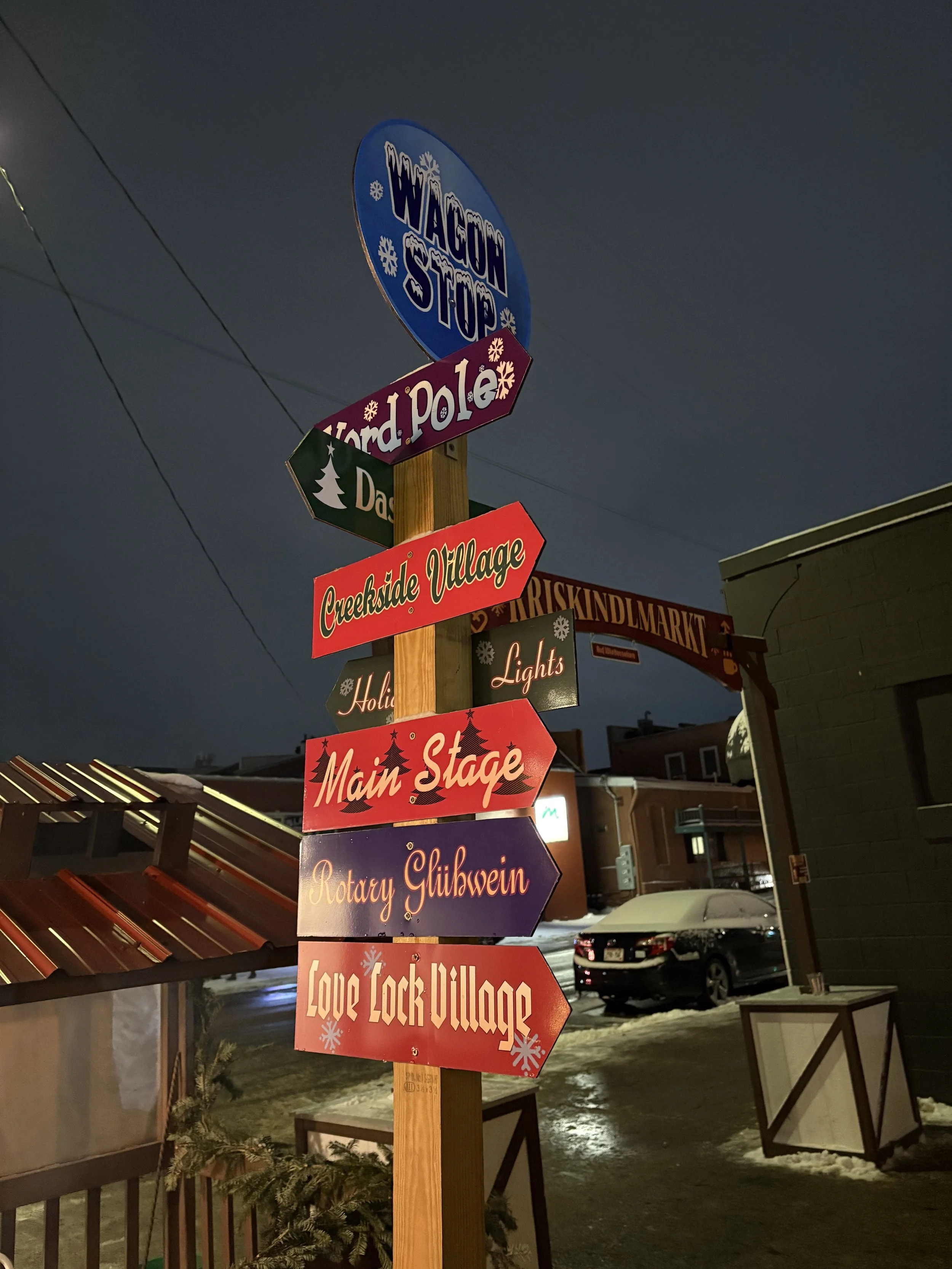 Colorful street signs with winter and holiday decorations, pointing to various locations including 'Dark Pole,' 'Creekside Village,' 'Main Stage,' and 'Love Lock Village,' near buildings and cars at night.