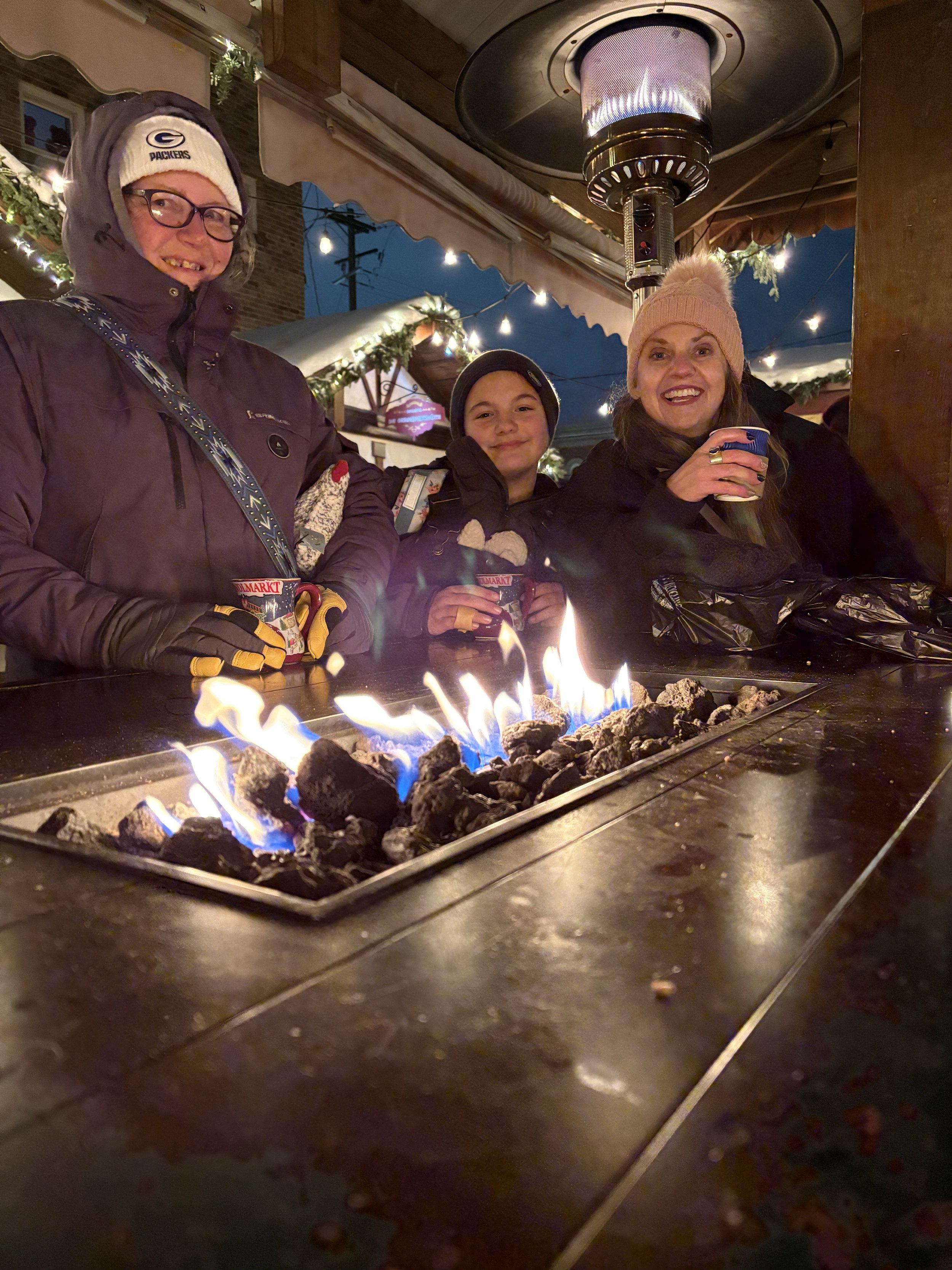 Three people sitting around a table with a fire pit in an outdoor restaurant at night, smiling and enjoying drinks, with festive string lights overhead and a heater above them.