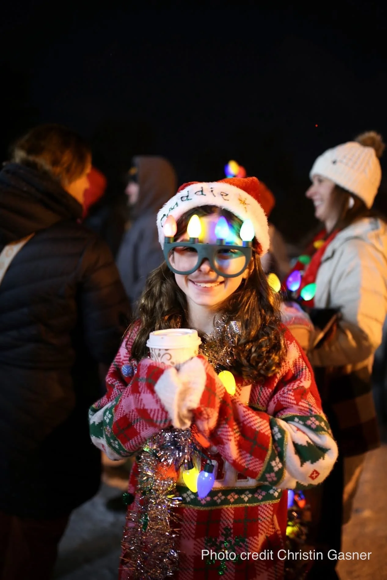 A young woman wearing Christmas lights on her head, a Santa hat, and festive sweater, smiling and holding a cup, at night during a holiday gathering.