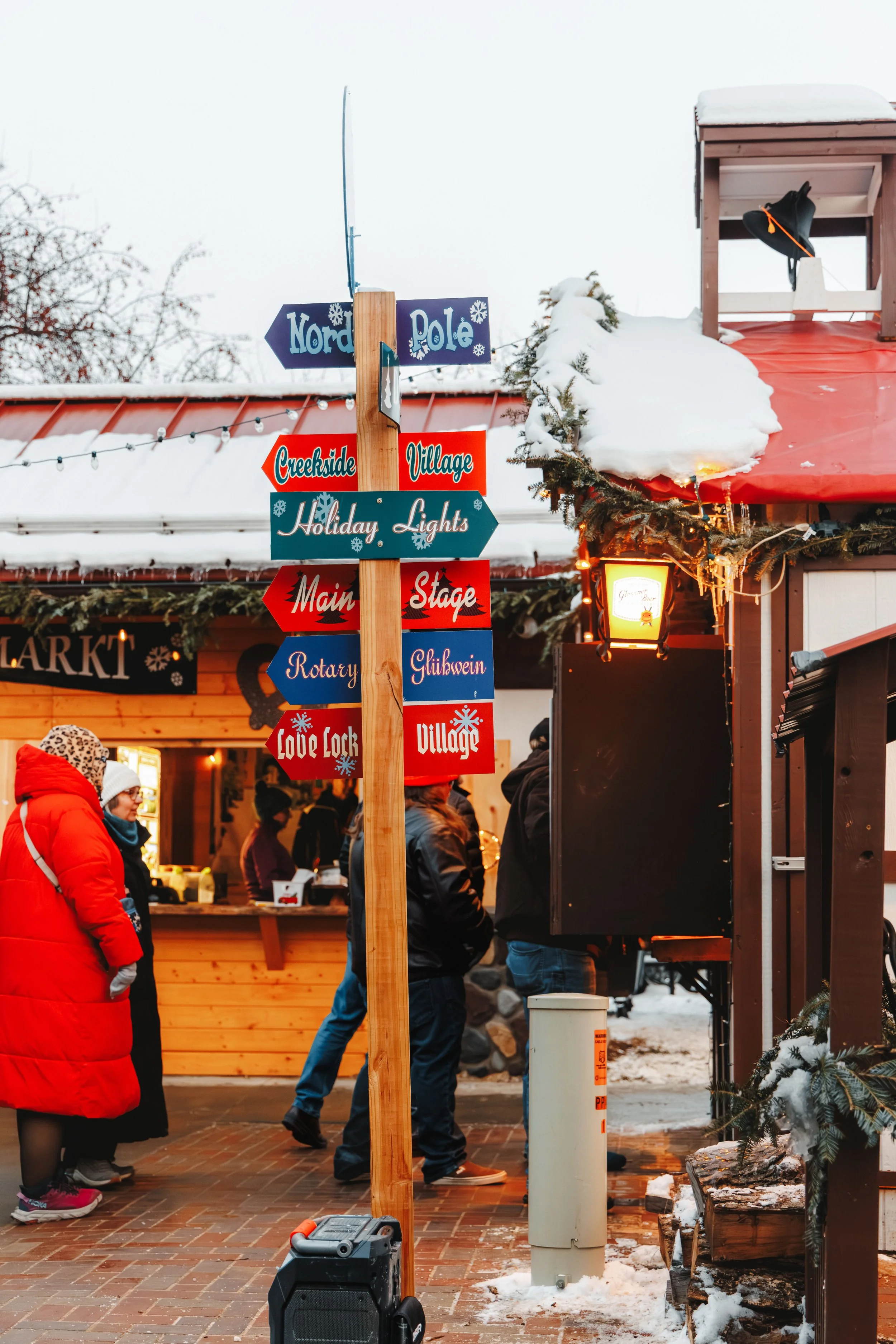 People standing near a wooden signpost with holiday and Christmas-themed signs, in front of wooden market stalls decorated with snow and evergreen branches, during a winter outdoor market.