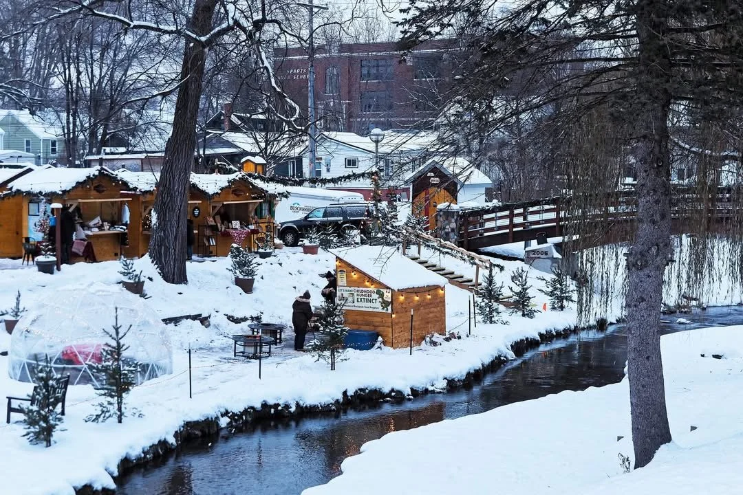 Snow-covered outdoor Christmas market near a creek, with small wooden stalls, string lights, trees, and a few people walking around.