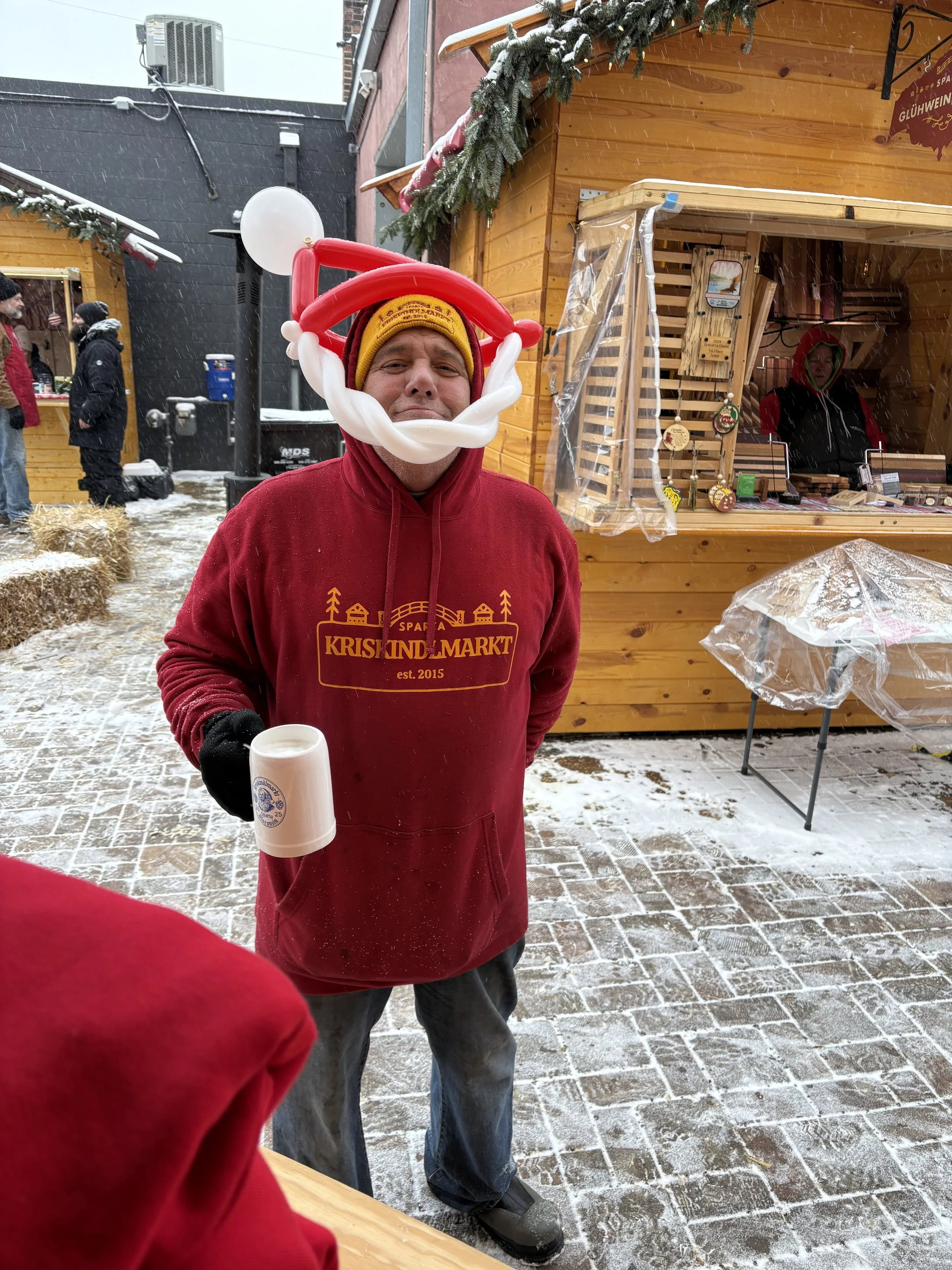 A man wearing a yellow beanie and red hoodie with gold text and images stands outdoors in snowy weather, wearing a boat-shaped balloon hat and holding a white mug. In the background, there is a wooden market stall decorated with evergreen branches and a woman inside.