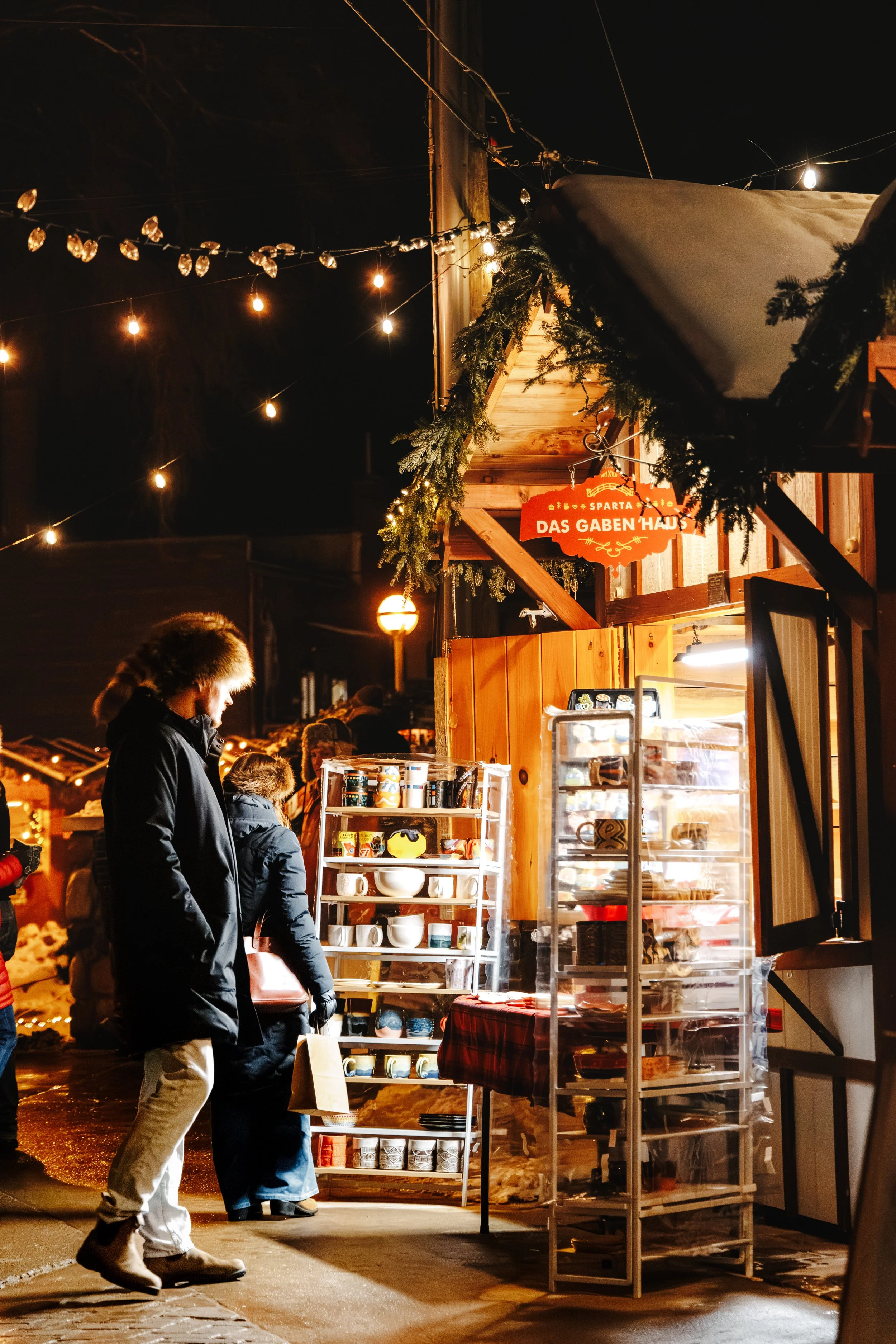 Night scene at a holiday market with a decorated wooden stall selling various ceramic mugs and dishes. String lights and greenery adorn the stall, and a red sign says