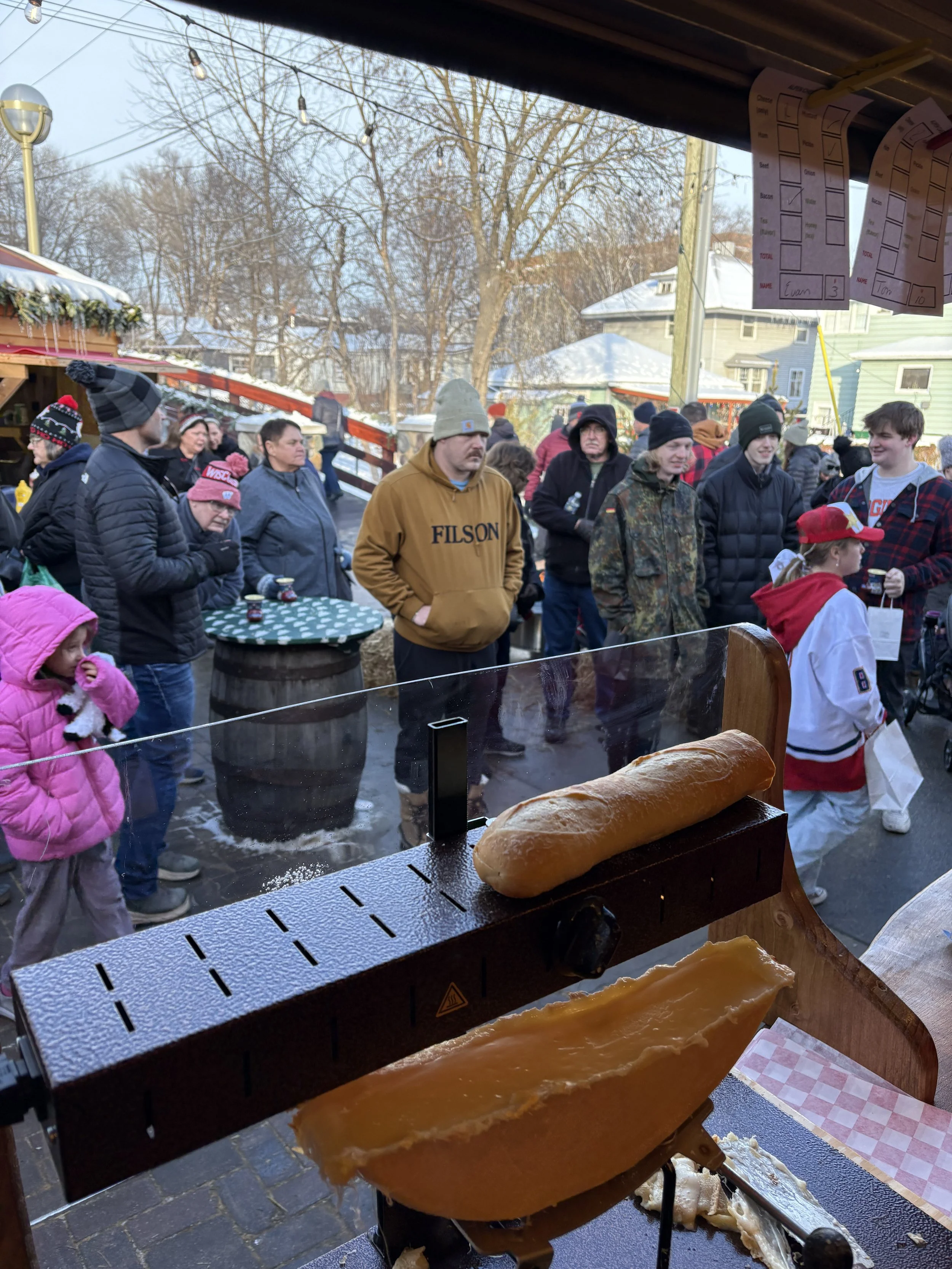 A crowd of people gathered outdoors in winter, with some wearing hats and jackets, near a food stand with a hot dog in the foreground.