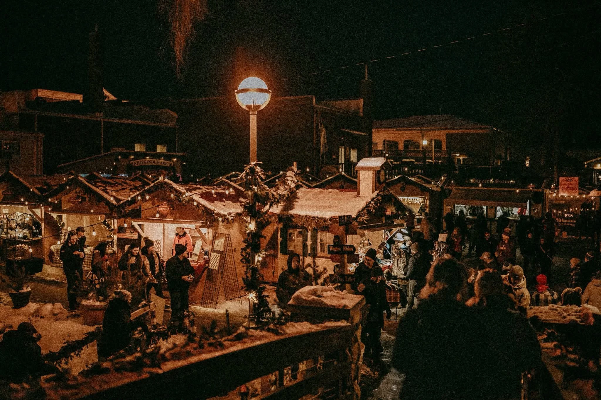 Nighttime scene at a festive outdoor market with people shopping and socializing, snow-covered rooftops, and string lights. A bright round moon or sun is seen in the dark sky.