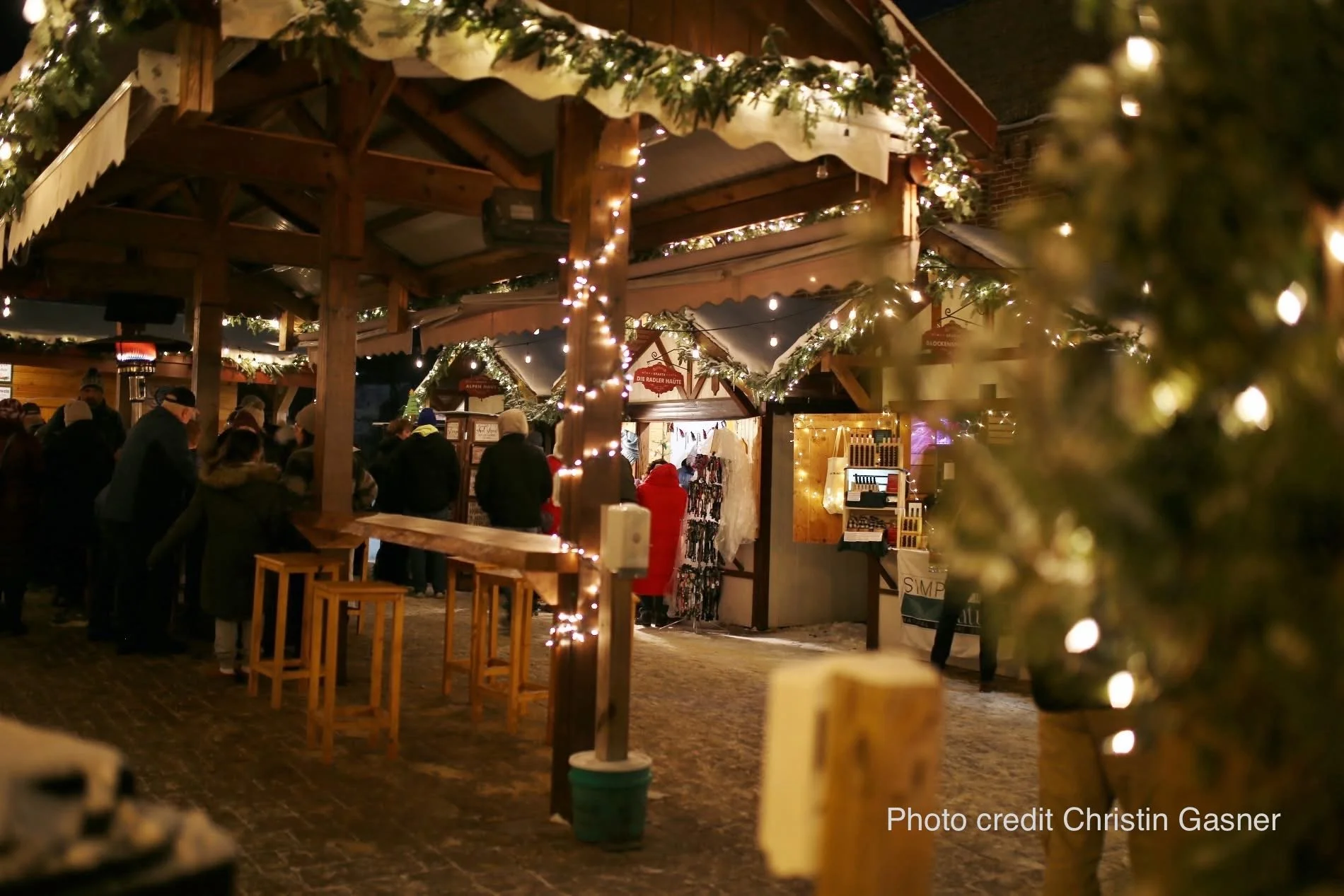 Nighttime holiday market with people gathered inside a decorated wooden structure adorned with string lights and greenery.