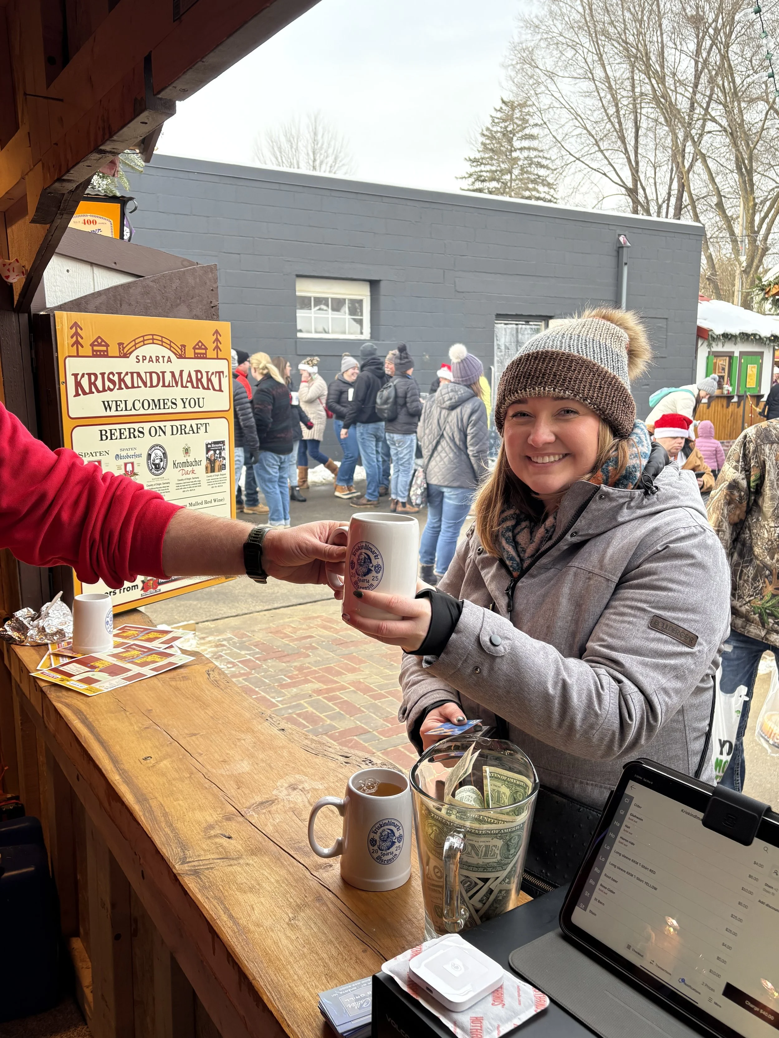A woman in a winter coat and knit hat smiles while holding a mug at an outdoor market. There is a sign that reads "Kriskindlmarkt" and various people in winter clothing in the background. Items like cash, a tablet, and beverages are on a wooden counter.