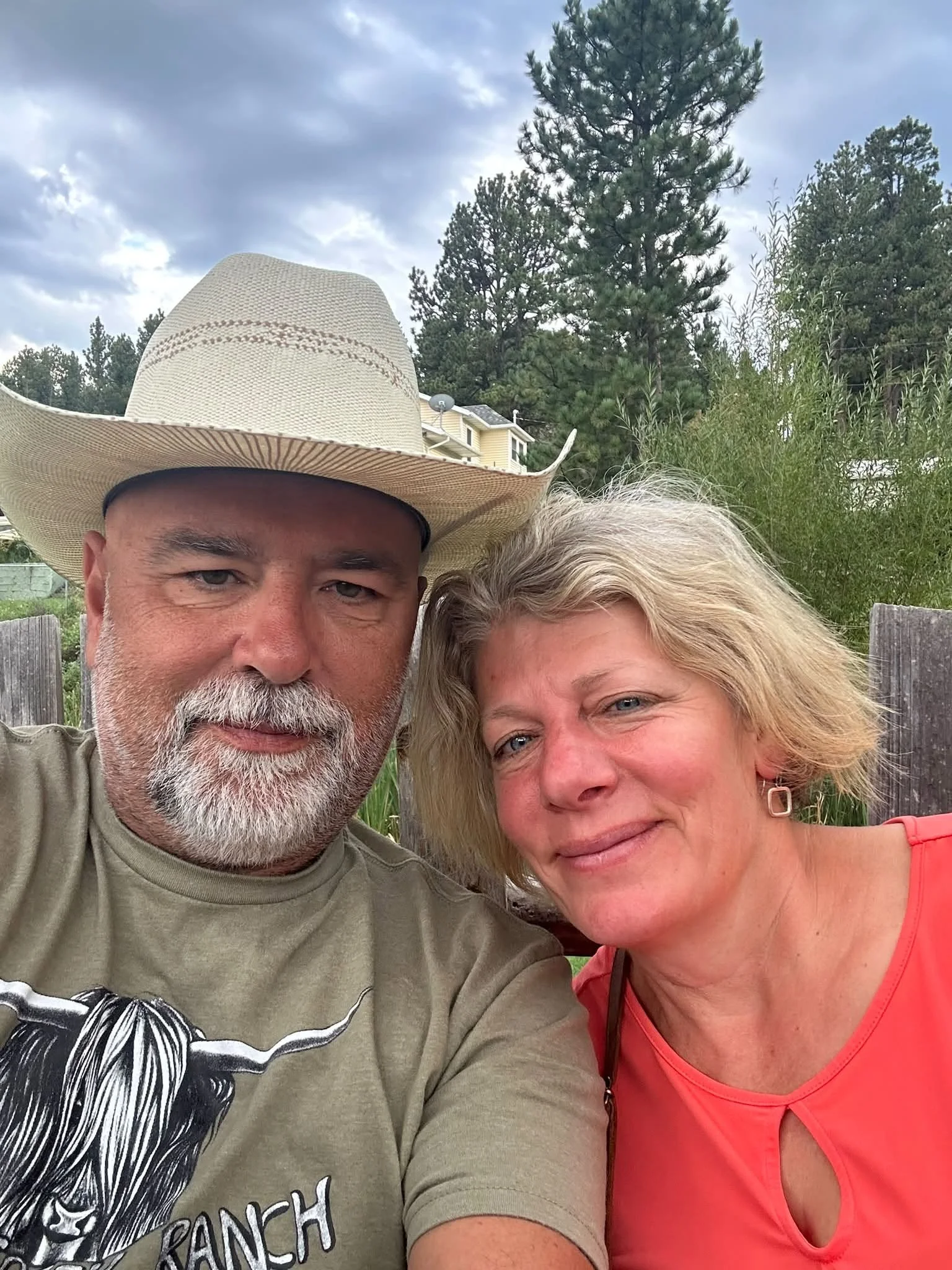 A middle-aged man with a beard and mustache wearing a large cowboy hat, and a middle-aged woman with blonde hair and blue eyes, smiling outdoors with trees and a cloudy sky in the background.