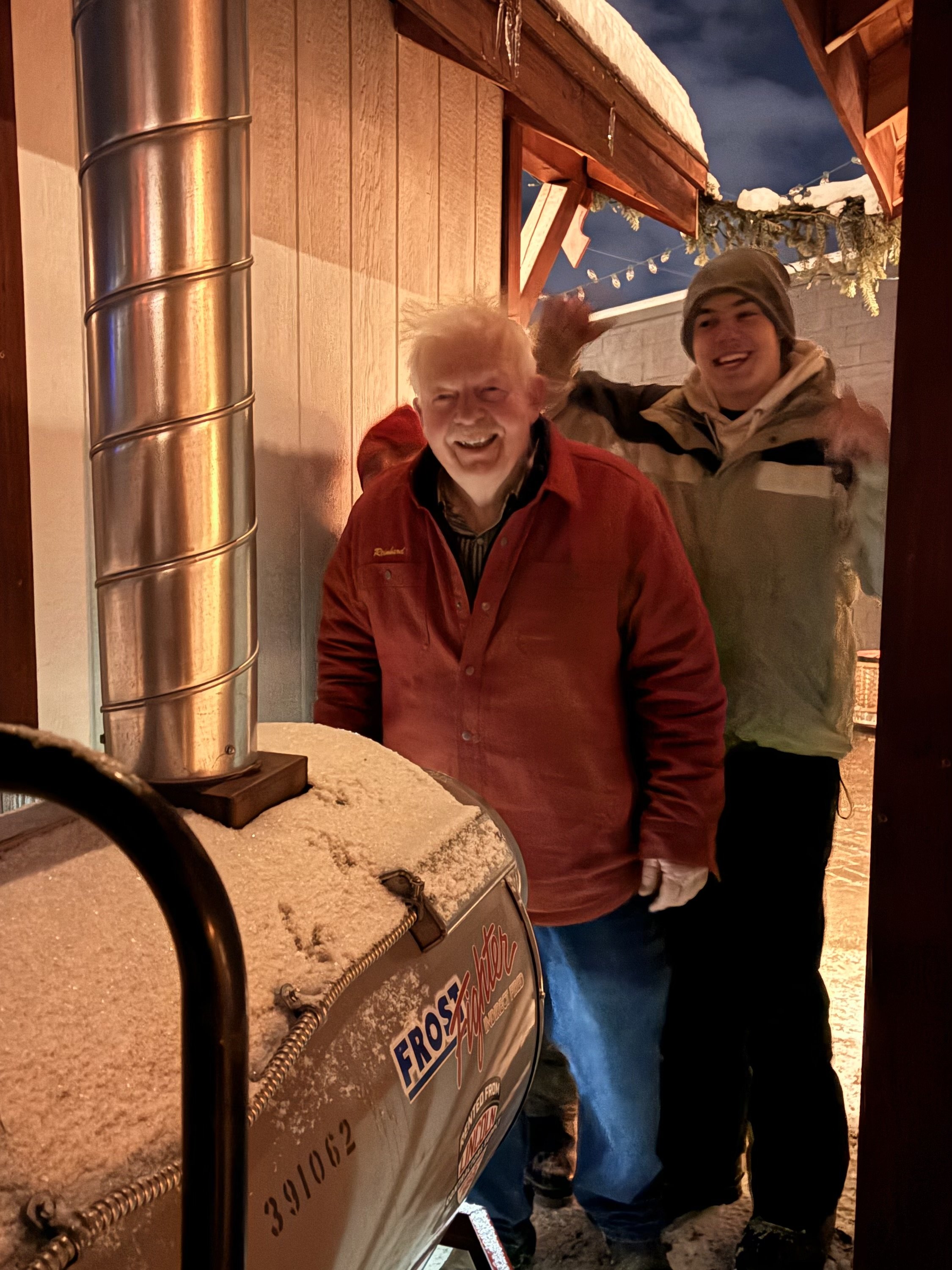 Two smiling men outdoors at night, one older with white hair and the other younger with a beanie, standing behind a snow-covered ice cream cart.