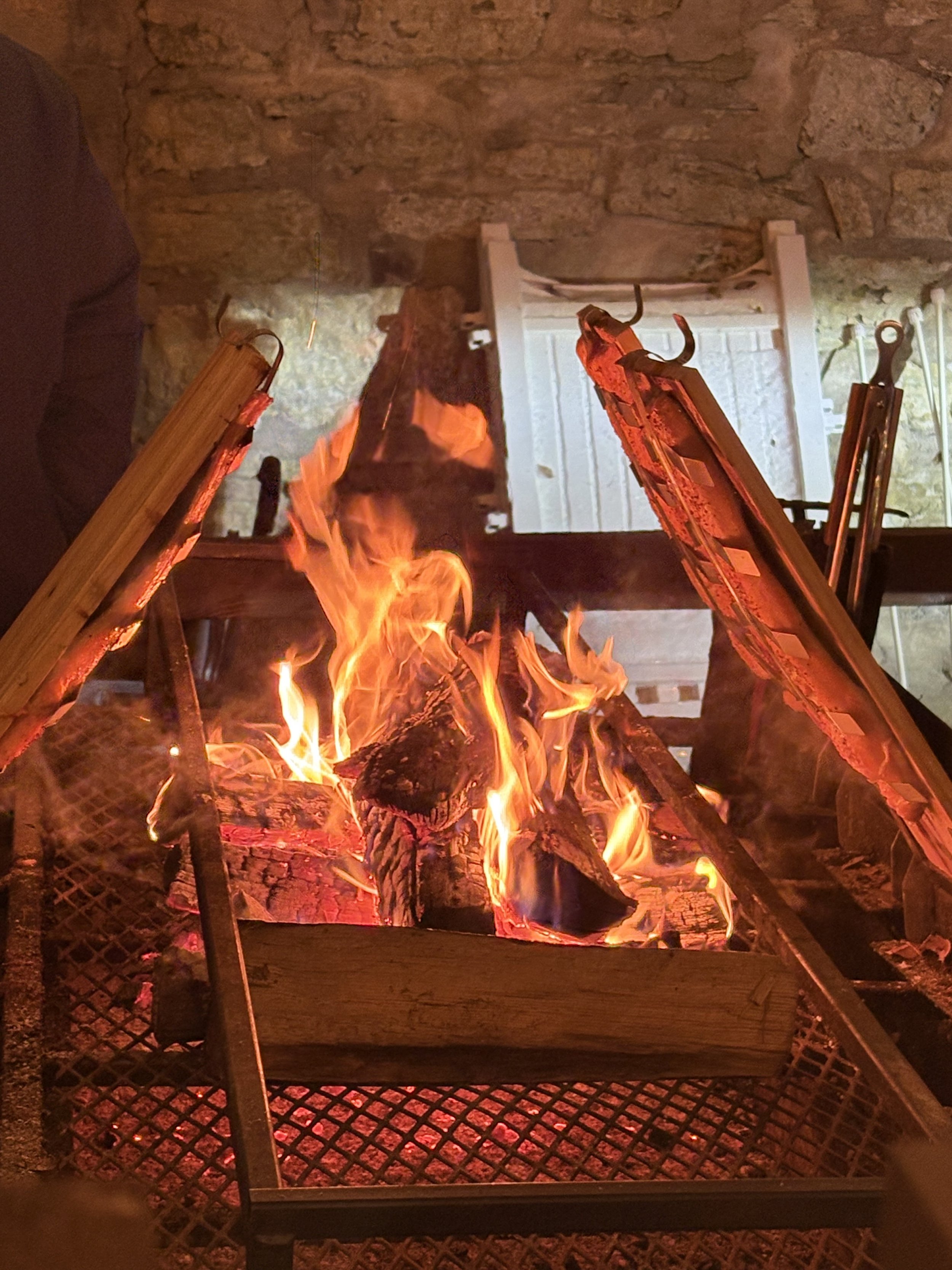 A small controlled outdoor fire with burning logs inside a metal fire pit. The fire is glowing with orange flames against a background of a stone or brick wall and part of a white outdoor porch or house. The fire is surrounded by a metal mesh or grate.
