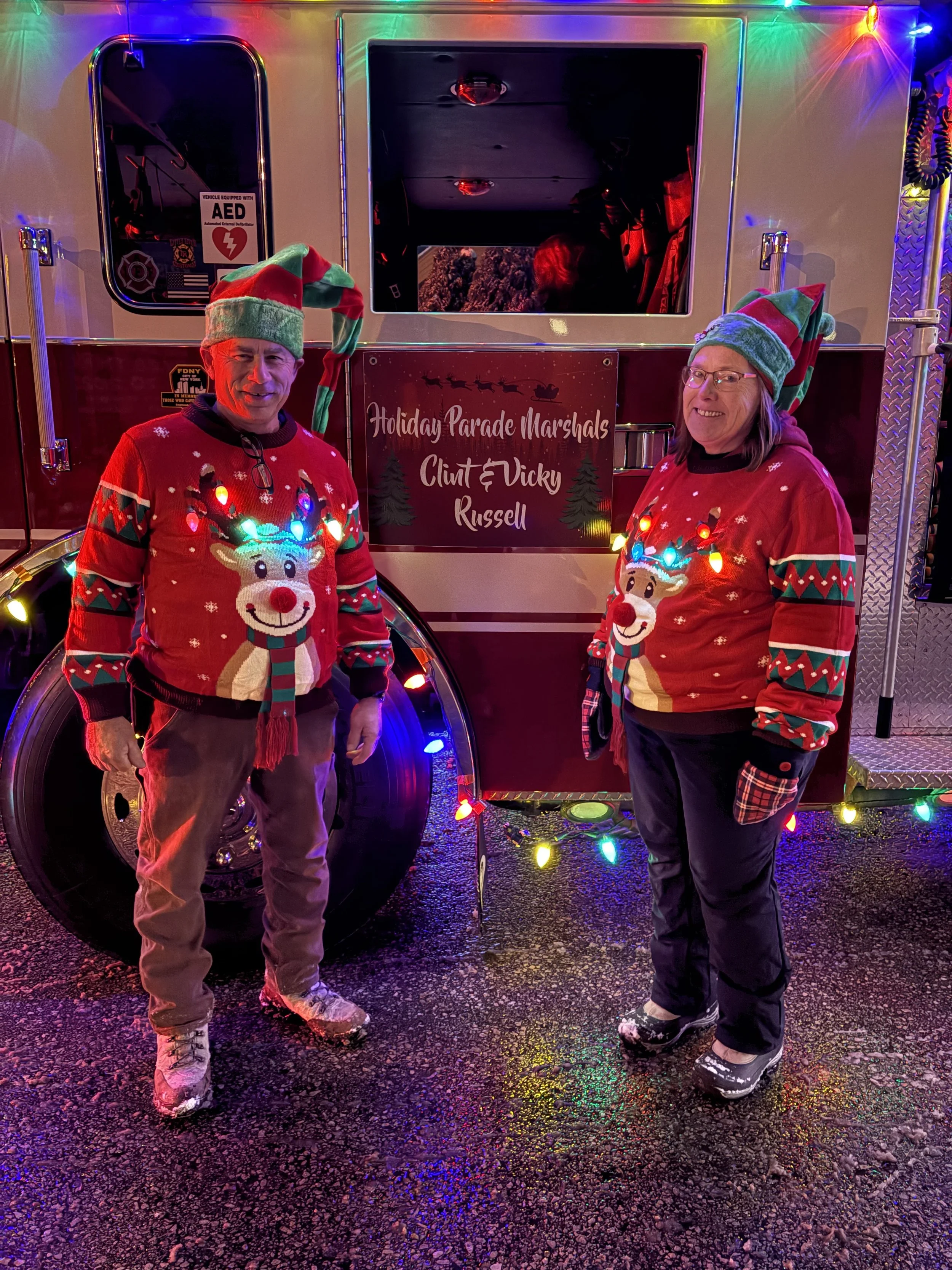 Two people in Christmas sweaters and elf hats standing in front of a decorated fire truck at night, with colorful string lights and a sign that reads 'Holiday Parade Marshals Clint & Vicky Russell'.