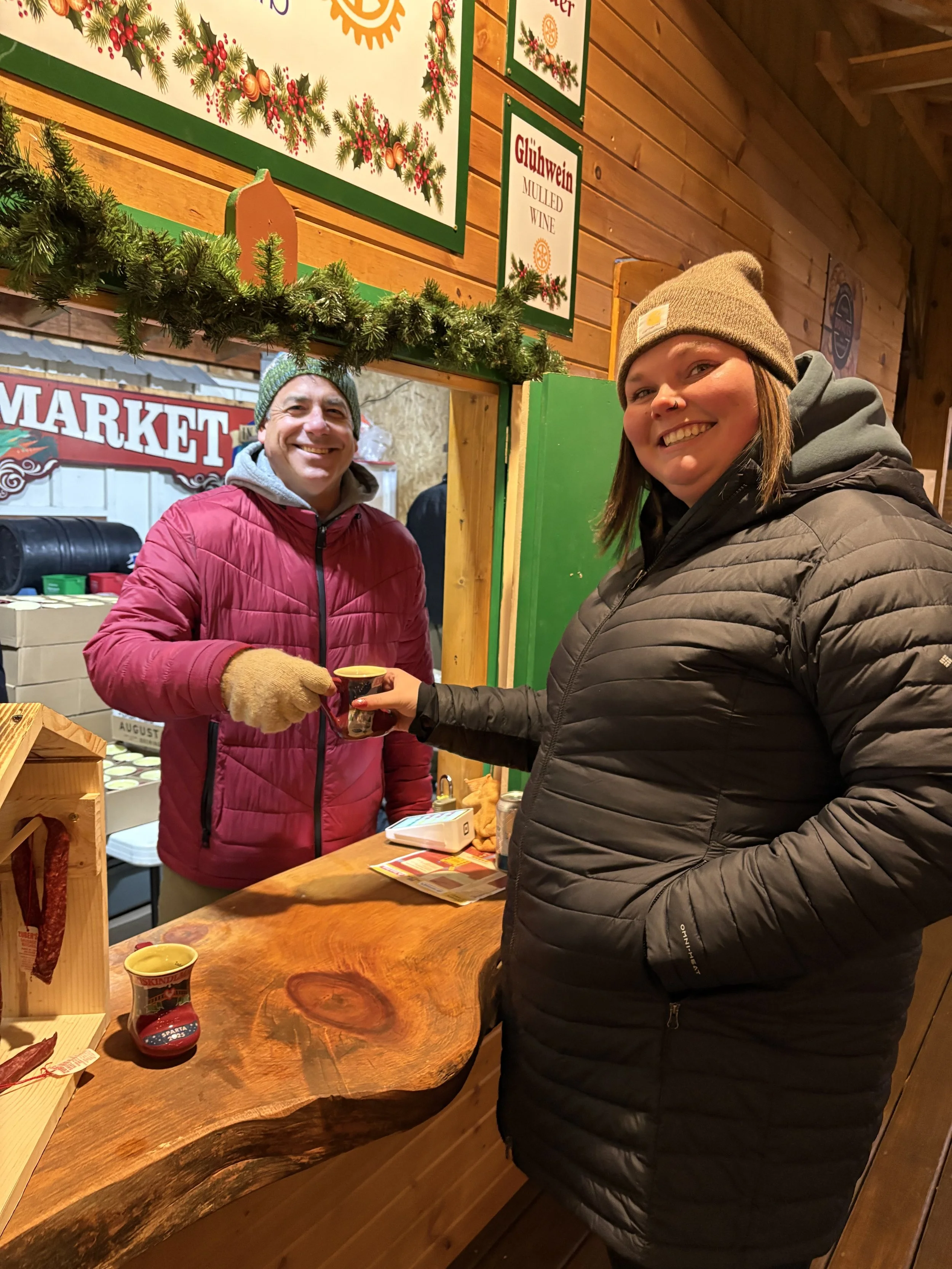 Two smiling people, a man and a woman, standing inside a wooden holiday market stall, with Christmas decorations and signs behind them. The woman is handing a small cup to the man. They are dressed warmly in jackets and hats.