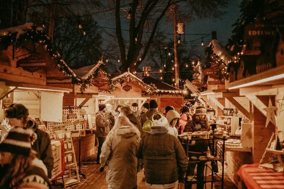 People shopping at a Christmas market stall decorated with string lights and garlands at night.