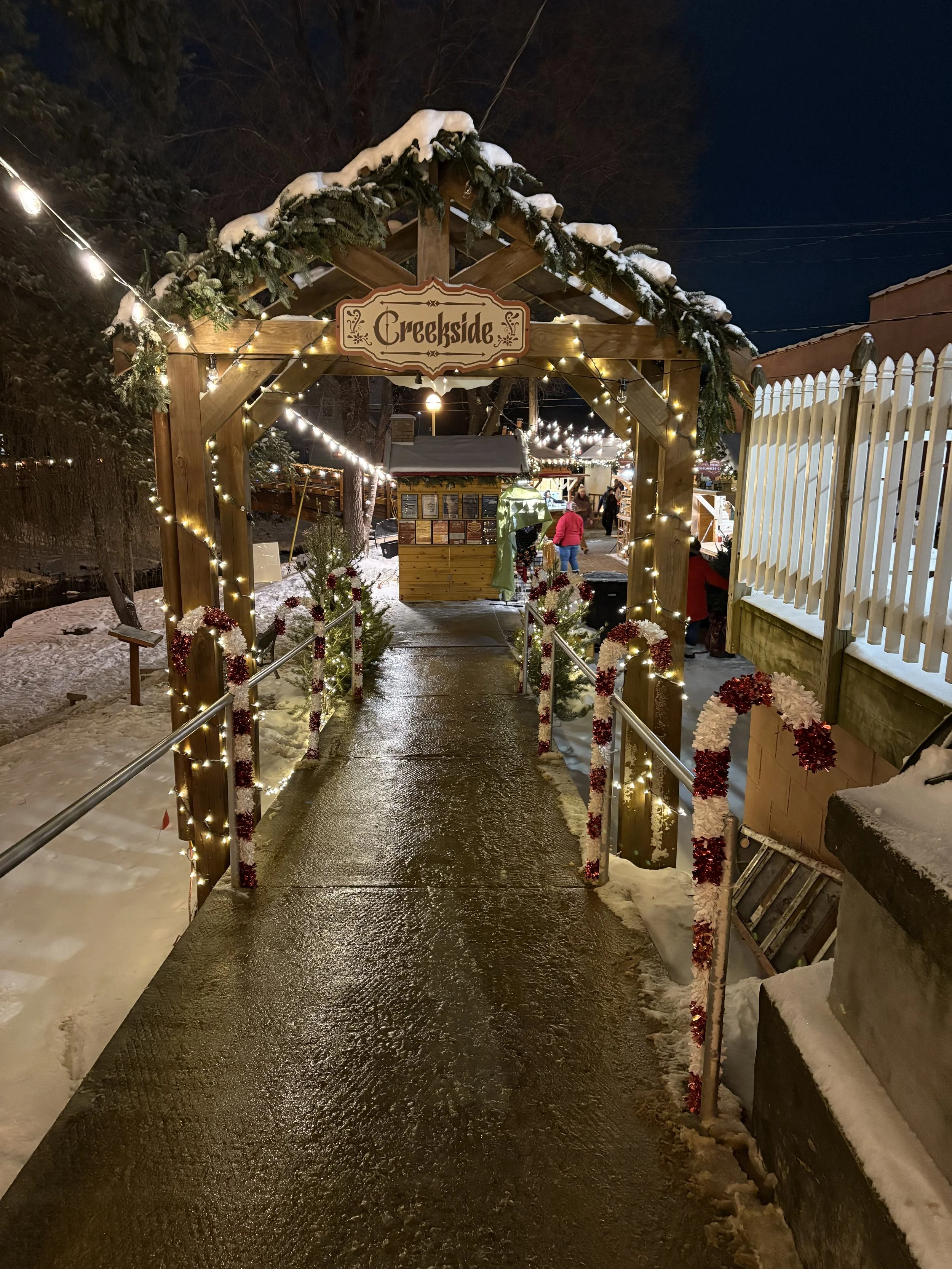 Entrance to a Christmas market called 'Creekside' decorated with string lights and garland, with snow on the ground and a small Christmas tree.