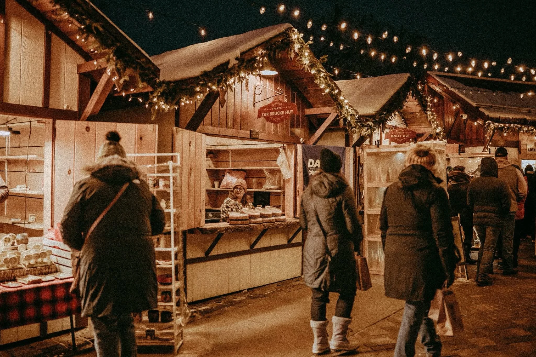 People shopping at a Christmas market stall decorated with string lights and garlands at night.