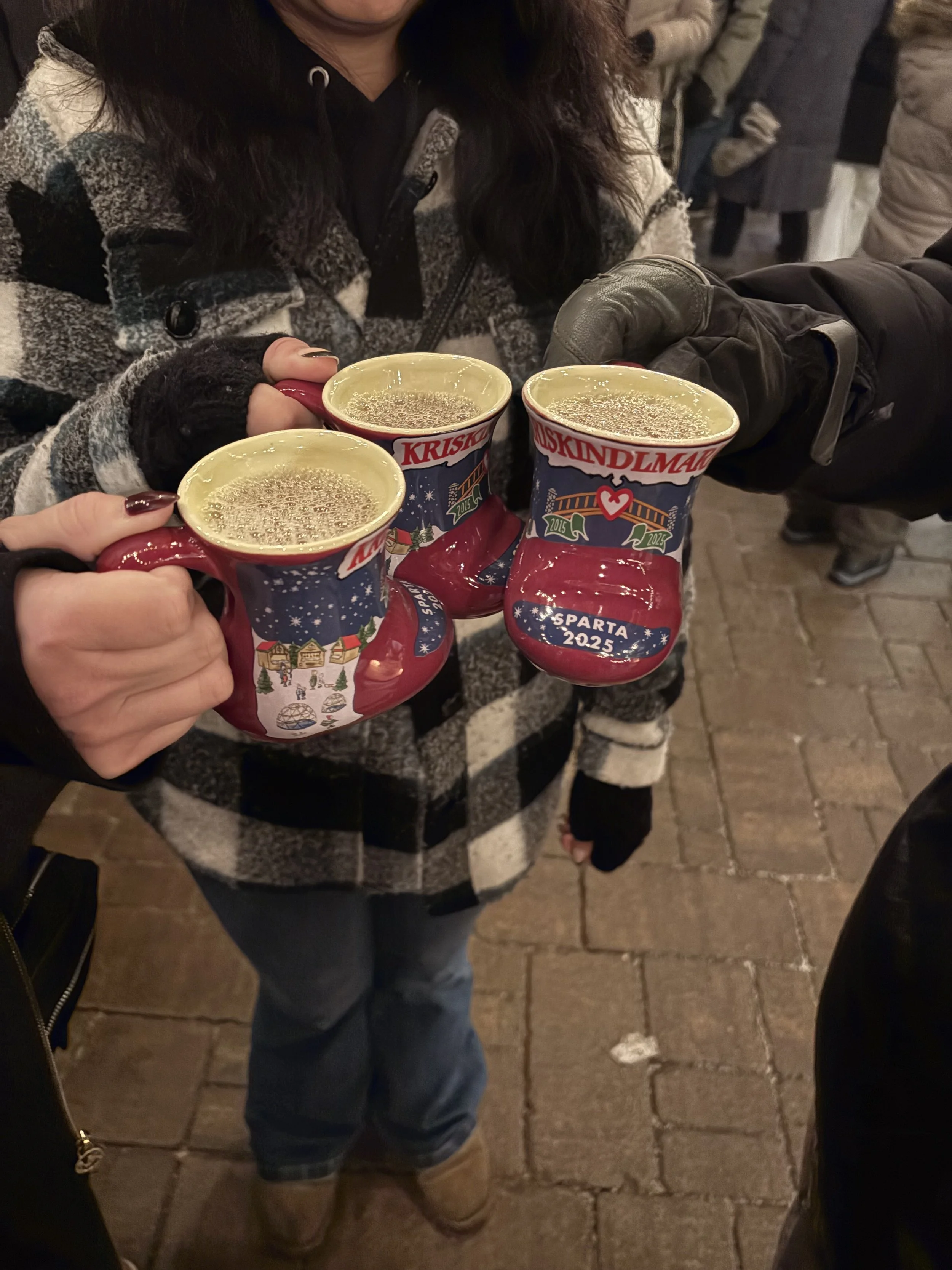 People gathering outdoors on a brick walkway, holding festive mugs with hot beverages, dressed in warm clothing for cold weather.