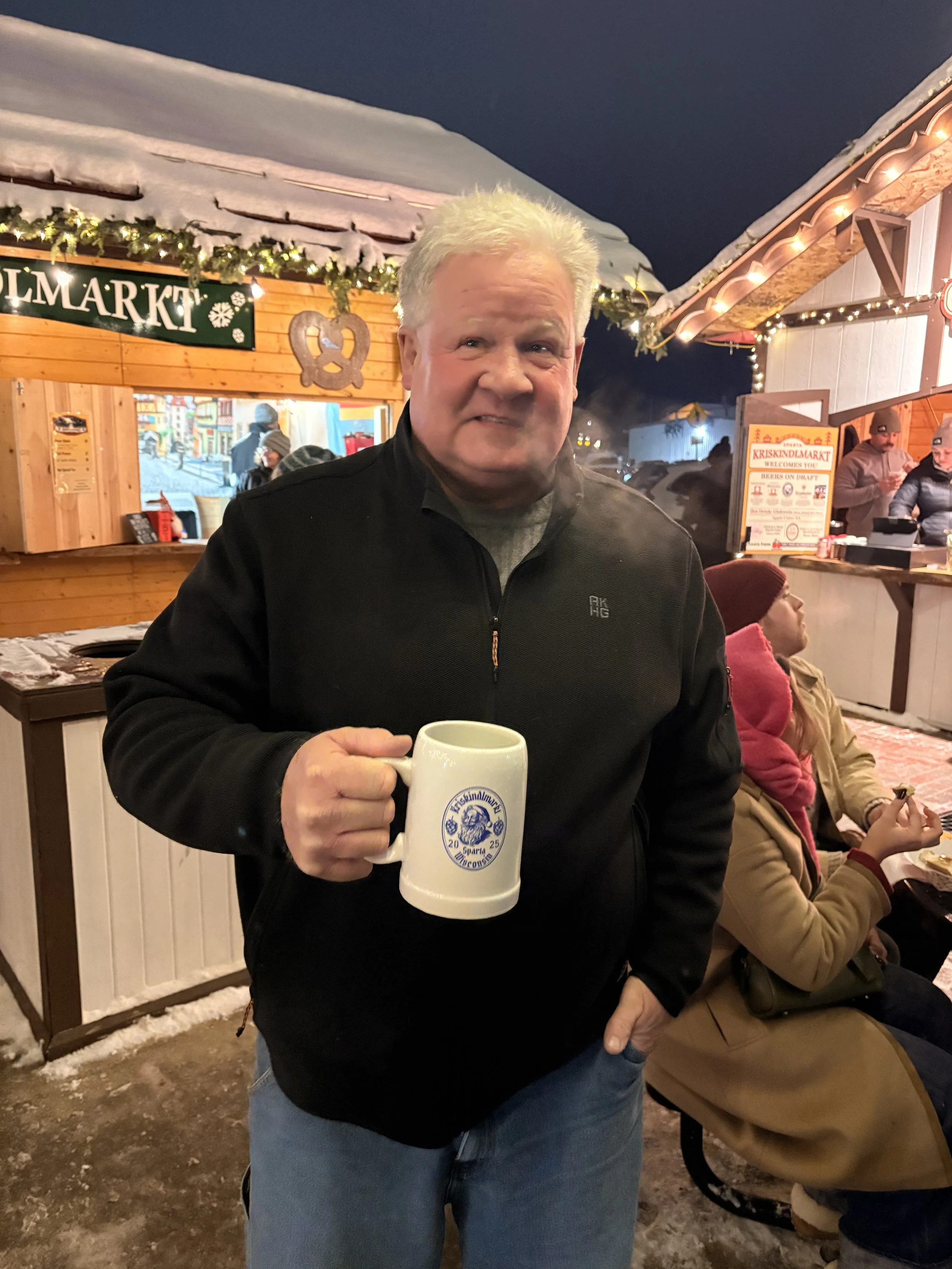A man with light hair wearing a black jacket holding a large white mug at a festive outdoor market during winter night. Snow covers the rooftops and ground, with Christmas lights and decorations visible in the background.