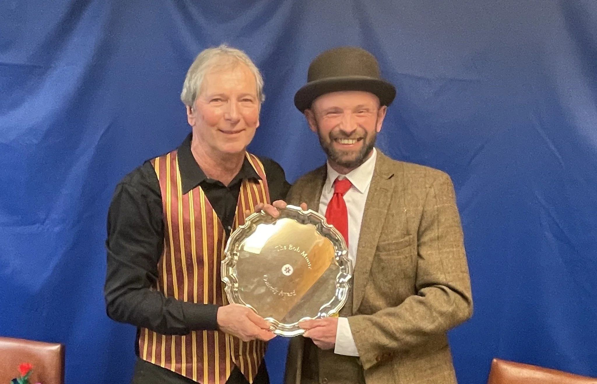 Two men smiling and holding a silver plate award in front of a blue backdrop. One man is in a black shirt and striped vest, the other is in a brown suit, white shirt, red tie, and wearing a brown bowler hat.