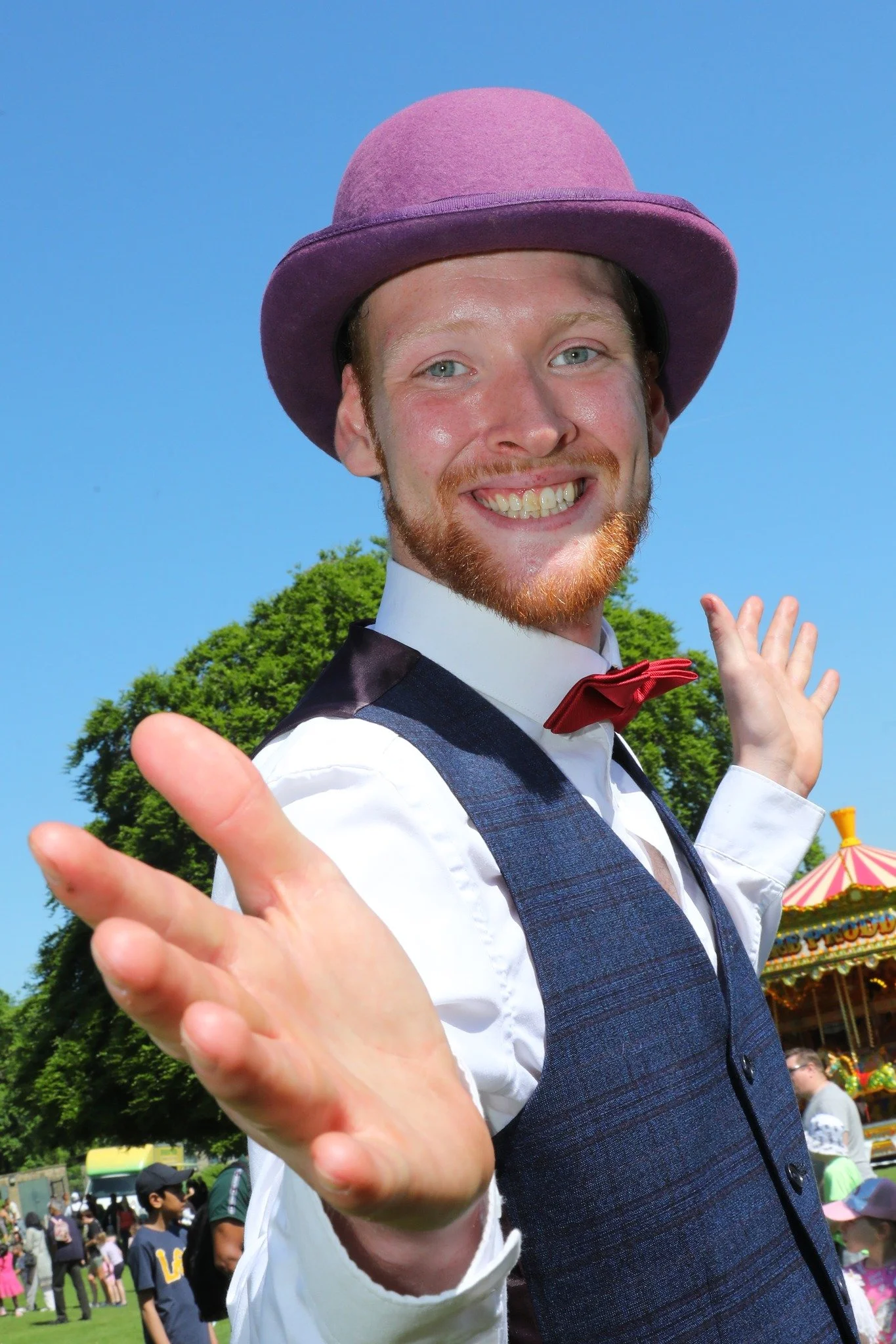 A man wearing a purple hat, red bow tie, white shirt, and vest, smiling and reaching out with both hands, in front of a background with trees and a carousel at an outdoor fair.