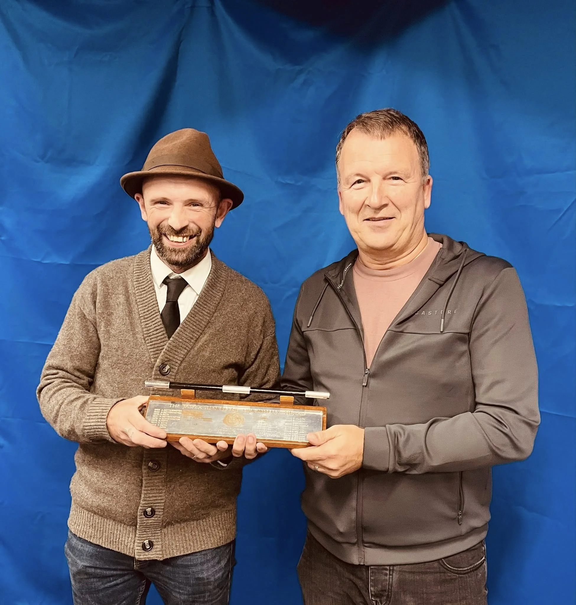 Two men standing side by side, holding a rectangular box with a pen on top, in front of a blue backdrop. The man on the left wears a brown hat, a brown cardigan, a white shirt, and a black tie; the man on the right wears a gray jacket over a pink shirt.