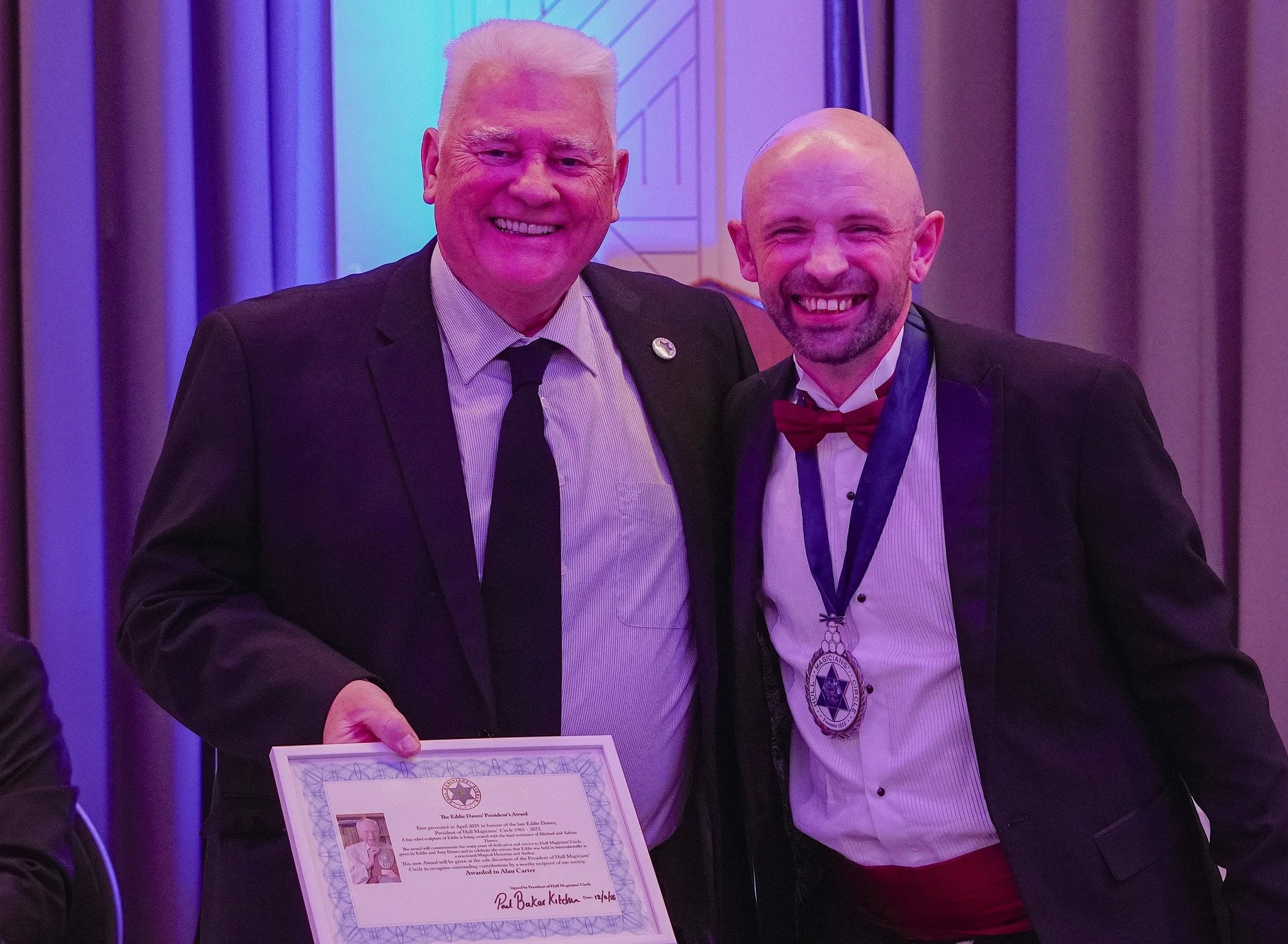 Two men in formal tuxedos smiling at a ceremony, one holding a certificate and the other wearing a medal around his neck.