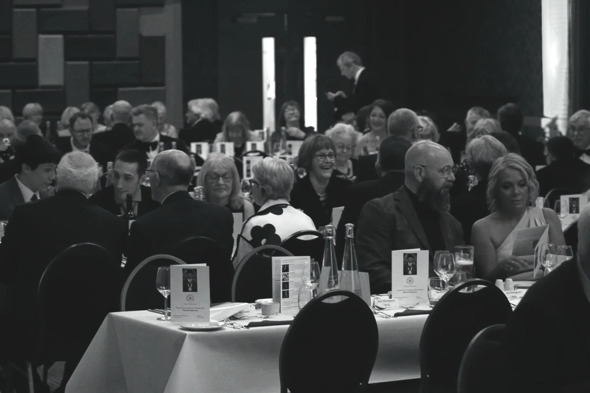 People seated at a formal dining event, dressed in evening attire, with table settings including wine glasses, bottles, and menus, in a large banquet hall.