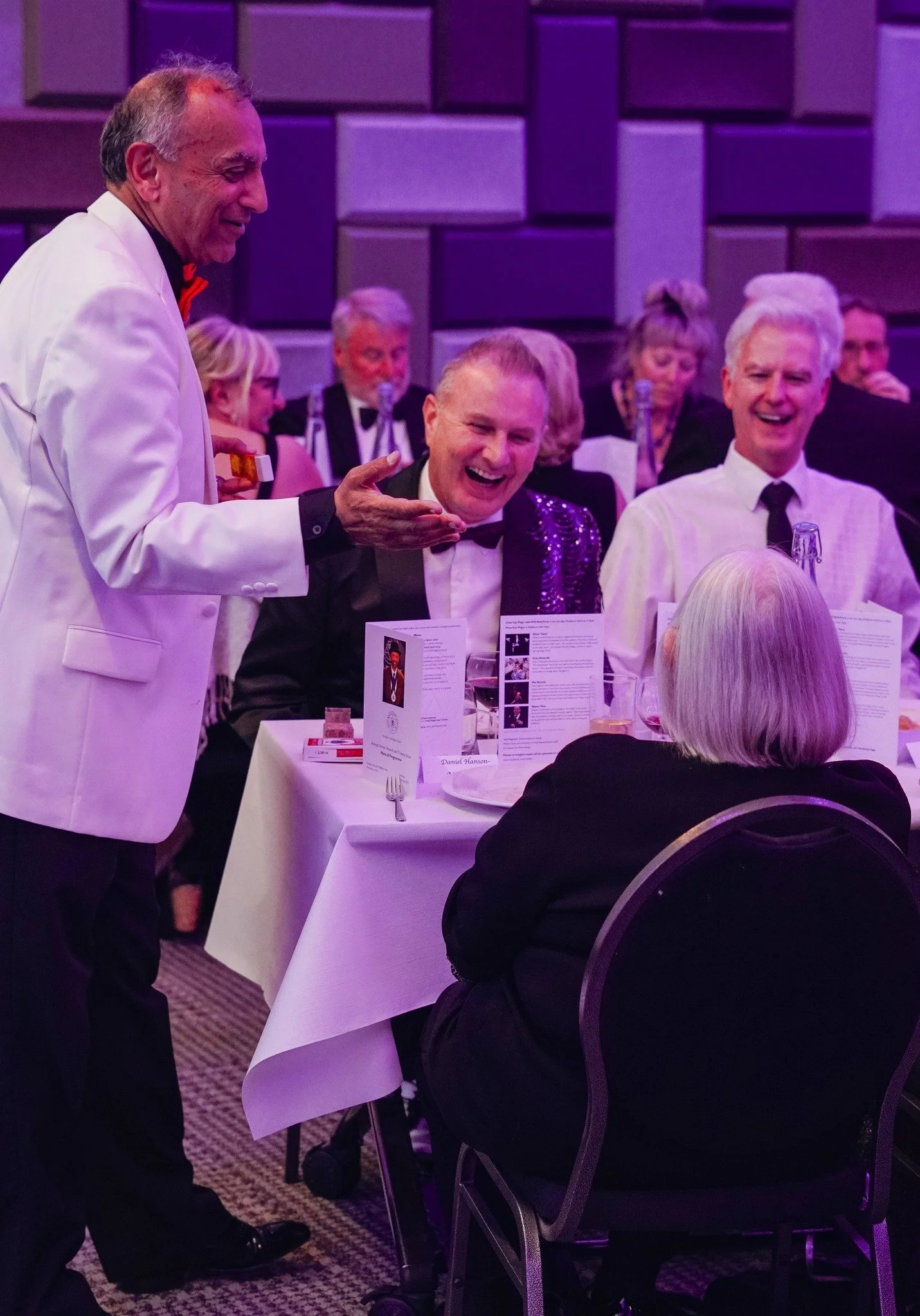 At a formal event, a waiter in a white jacket is serving a group of people sitting at a table, with purple lighting and a decorated background.