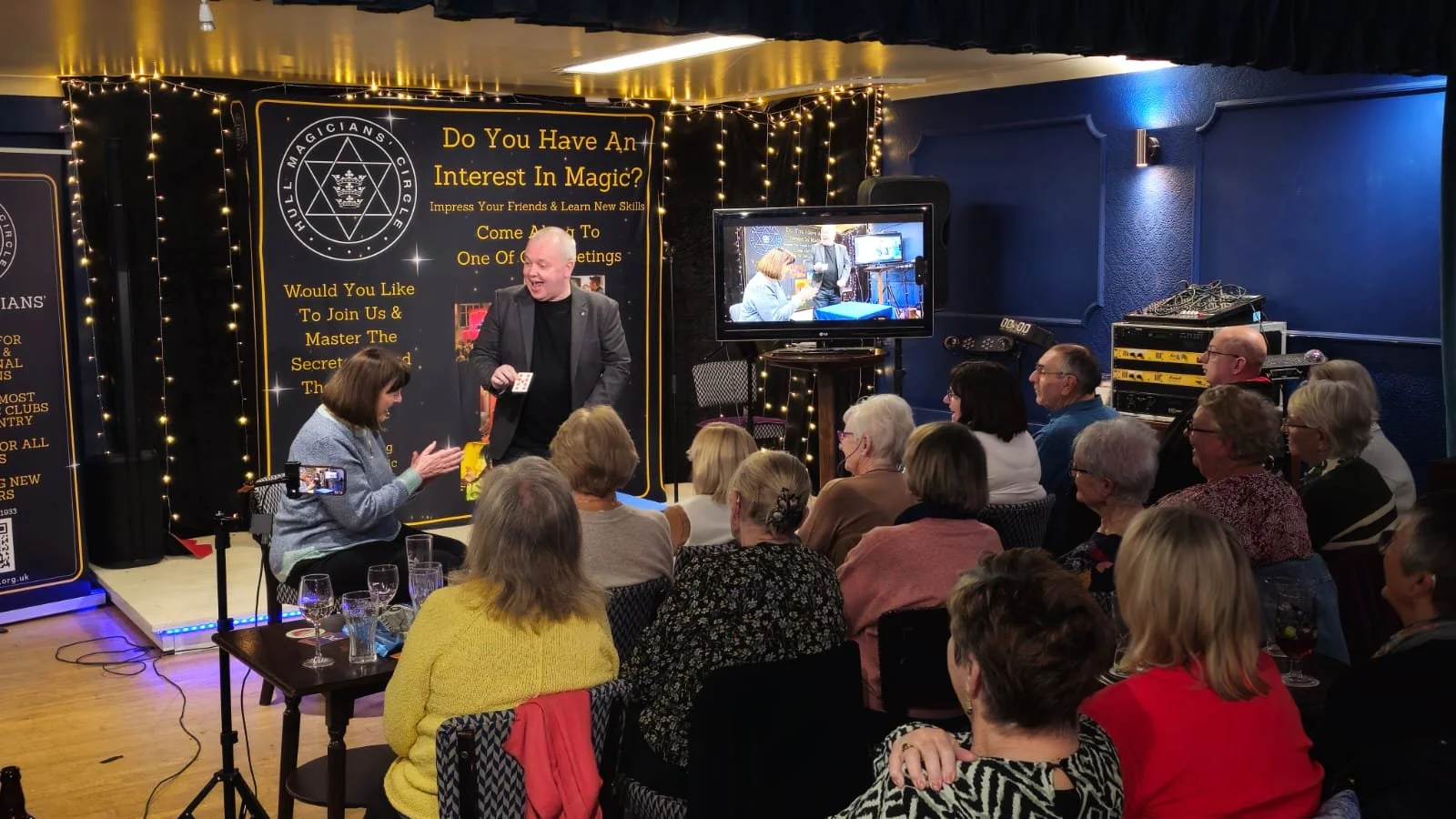A magic show performer in a black suit entertains an audience of mostly women in a room decorated with fairy lights. A woman in the front row appears to be reacting to the performance, with a phone on a tripod recording. There is a screen showing the