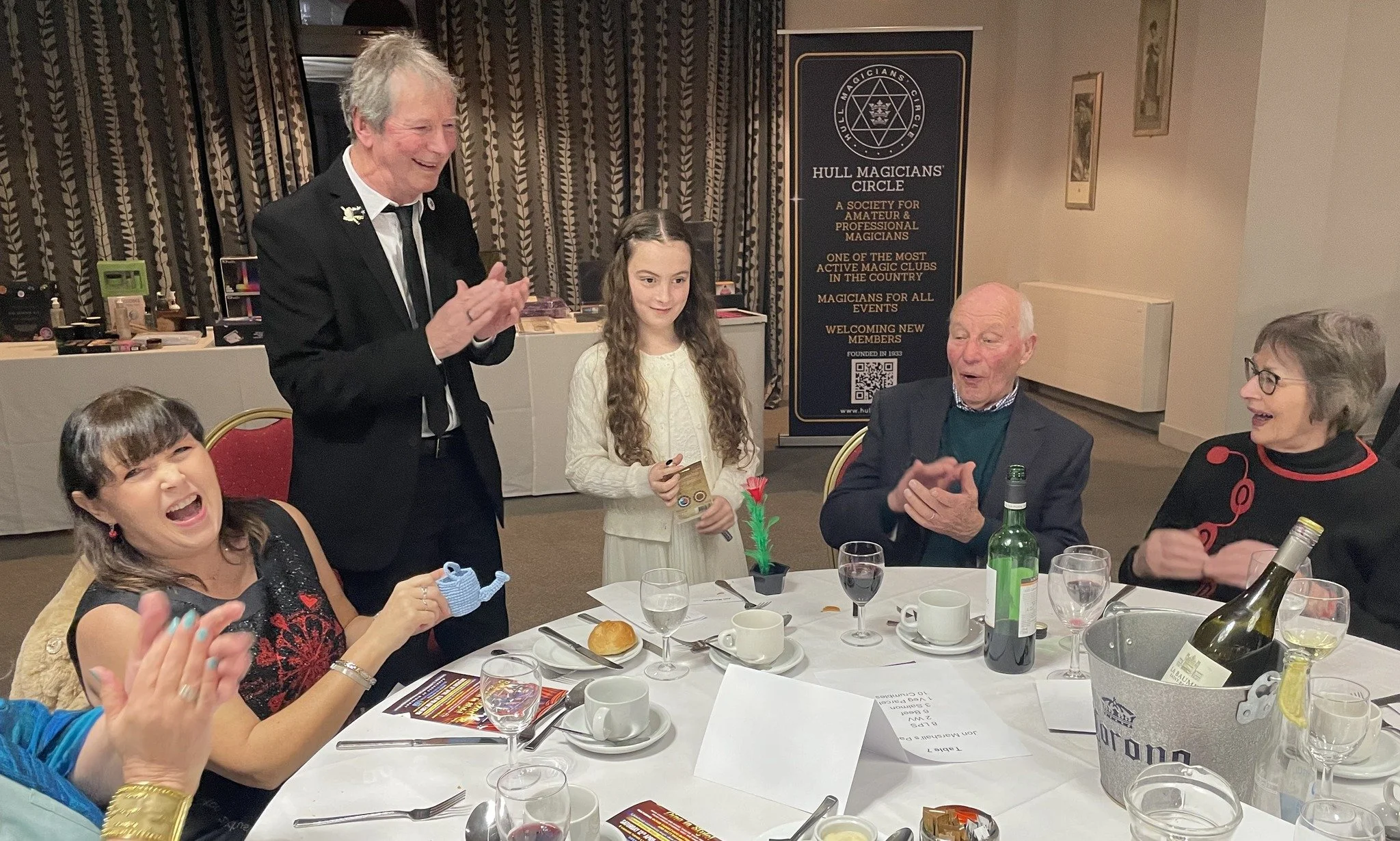 Group of people celebrating at a dinner event, with a young girl receiving a gift, laughing and clapping, in a banquet room with a banner for the Hull Magicians' Circle in the background.