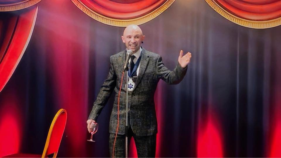 A man in a plaid suit with a microphone, standing on stage with red curtains and a dark backdrop, holding a glass of wine and smiling.