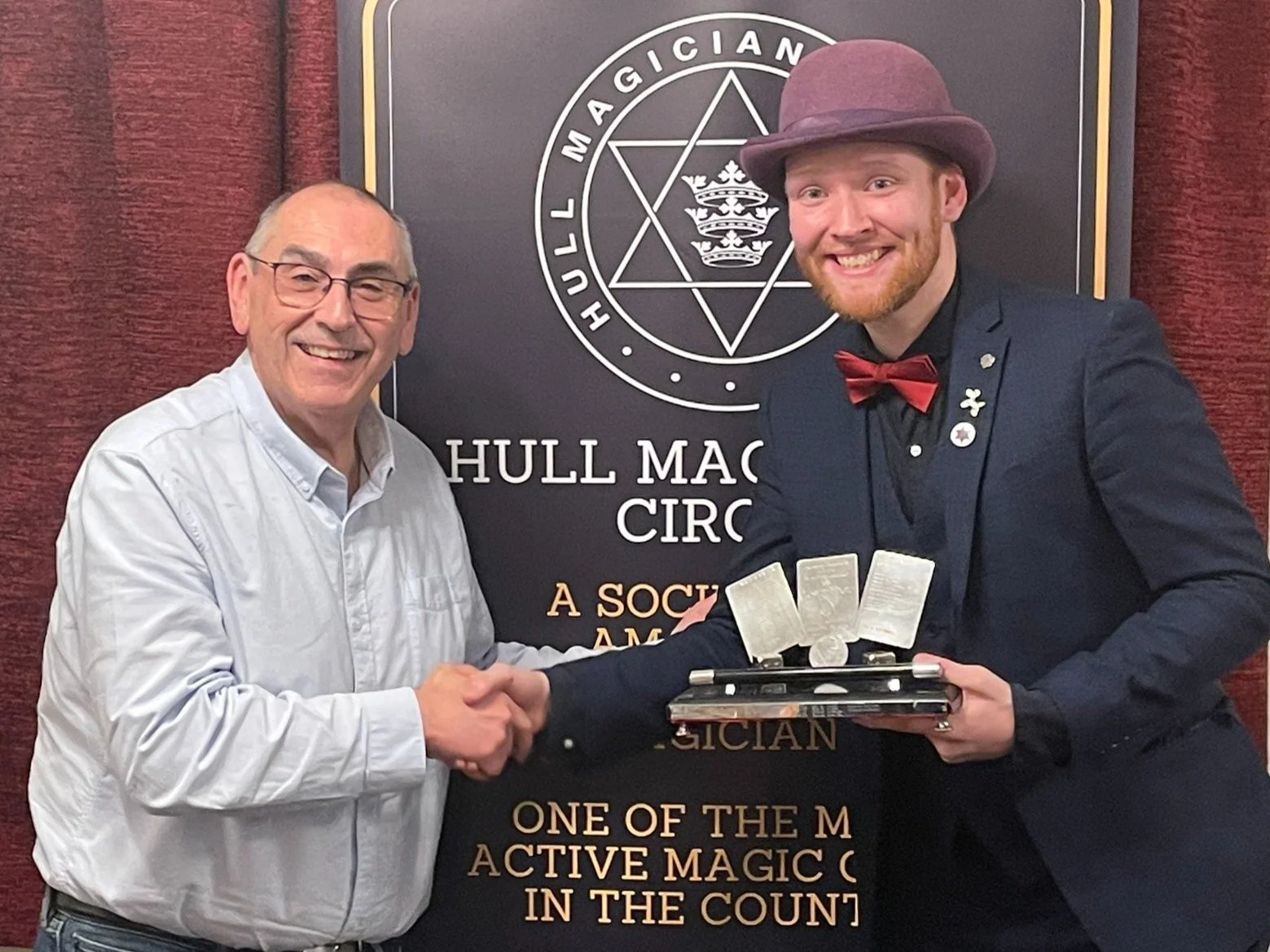 Two men shaking hands in front of a Hull Mac Circus banner. The man on the left is wearing glasses and a white shirt, smiling. The man on the right has red hair and a beard, wearing a dark suit with a red bow tie, a purple hat, and various pins on his lapel. He's holding a trophy with cards on top.