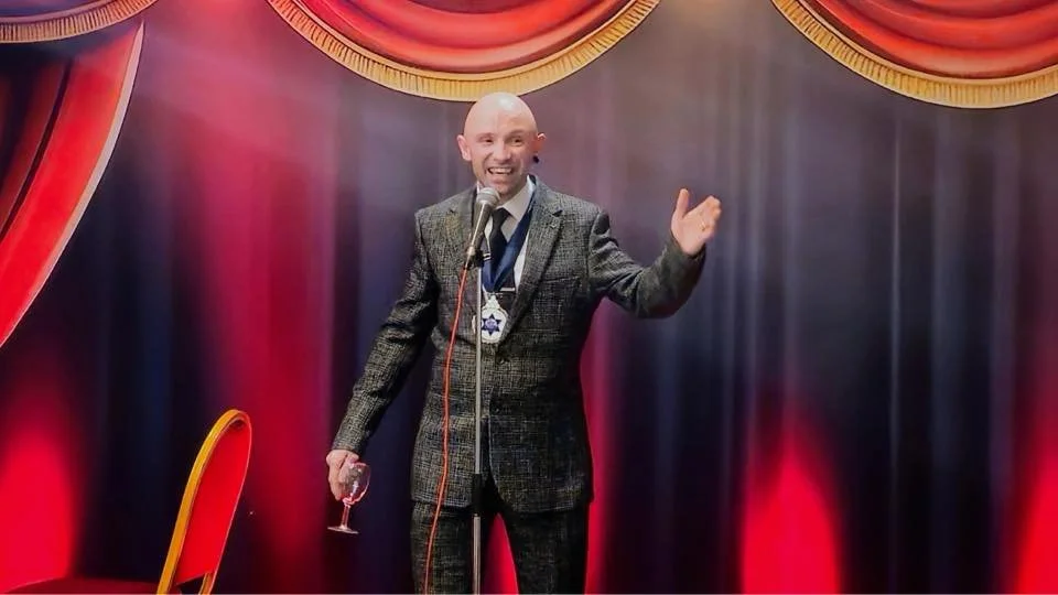 A man in a suit and tie, holding a glass of wine, standing on stage with a microphone, smiling and waving, with red curtains and stage lighting in the background.