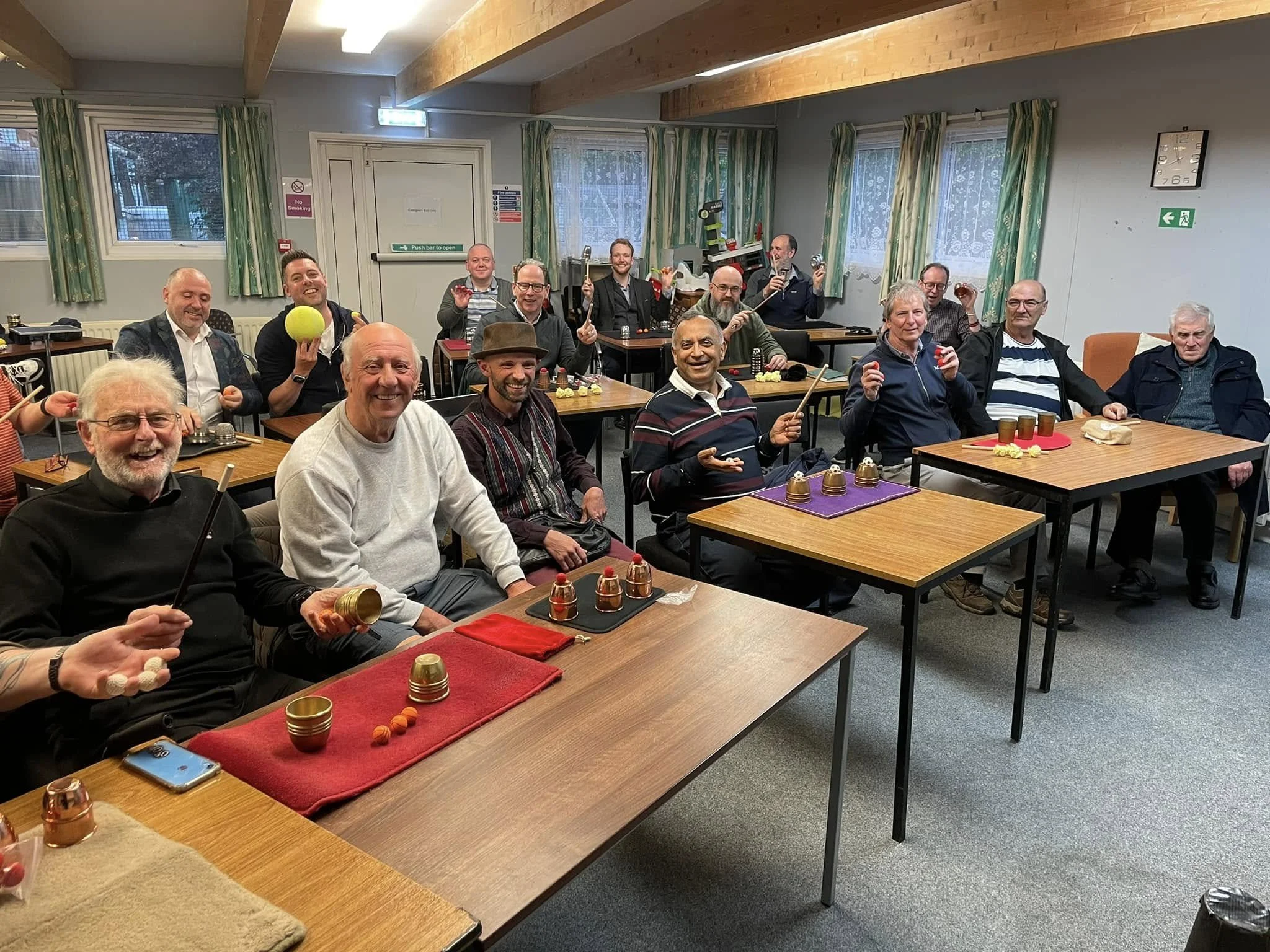 A group of smiling men gathered in a classroom or community room, sitting at tables with small bells, balls, and cups, holding musical instruments and small balls, appearing to be in a meeting or celebration.