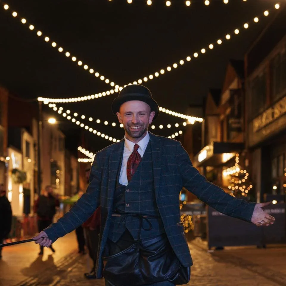 A man dressed in vintage attire, standing on a city street at night with string lights overhead, smiling and gesturing with his arms spread.