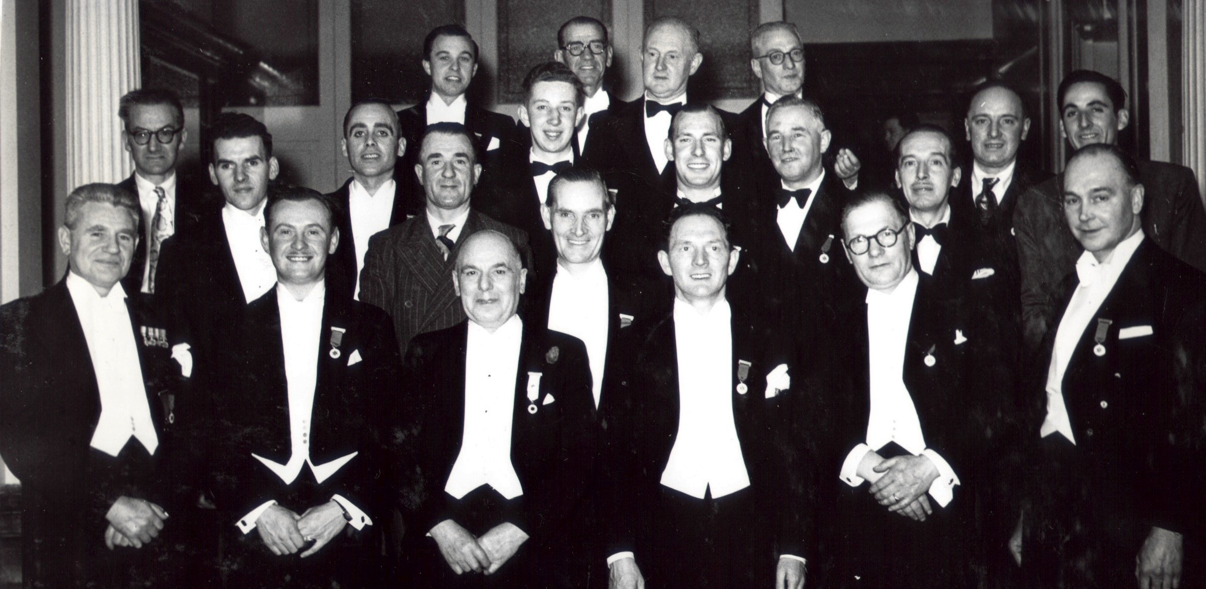 Black and white photograph of a group of men dressed in formal tuxedos, standing indoors. Some men are wearing medals on their jackets, and they are posed together for a group photo.