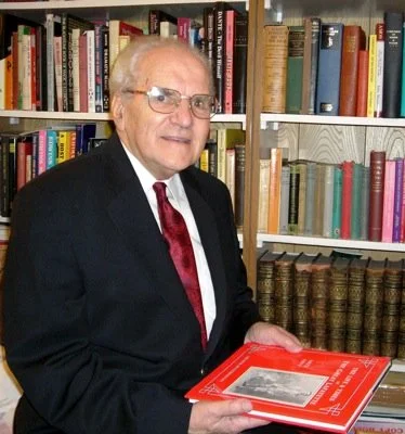 An elderly man in a suit and glasses standing in front of a bookshelf, holding a red book.