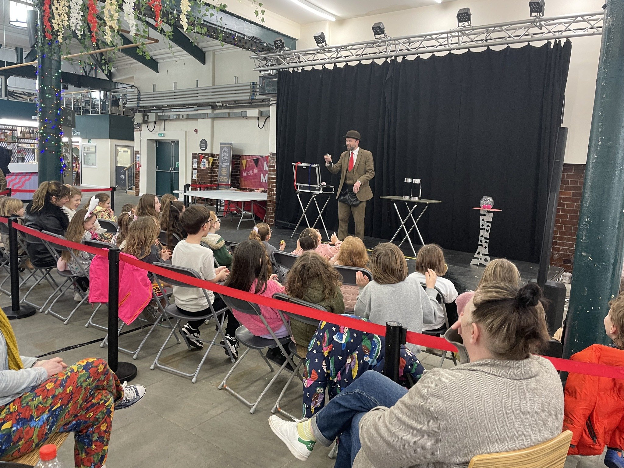 A magician performing on stage for a group of children sitting in chairs, with some wearing colorful clothing, in an indoor venue decorated with seasonal ornaments and string lights.