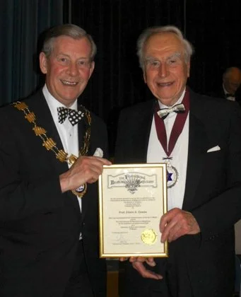 Two men in tuxedos, one wearing a mayor's chain, holding a framed certificate at an awards ceremony.