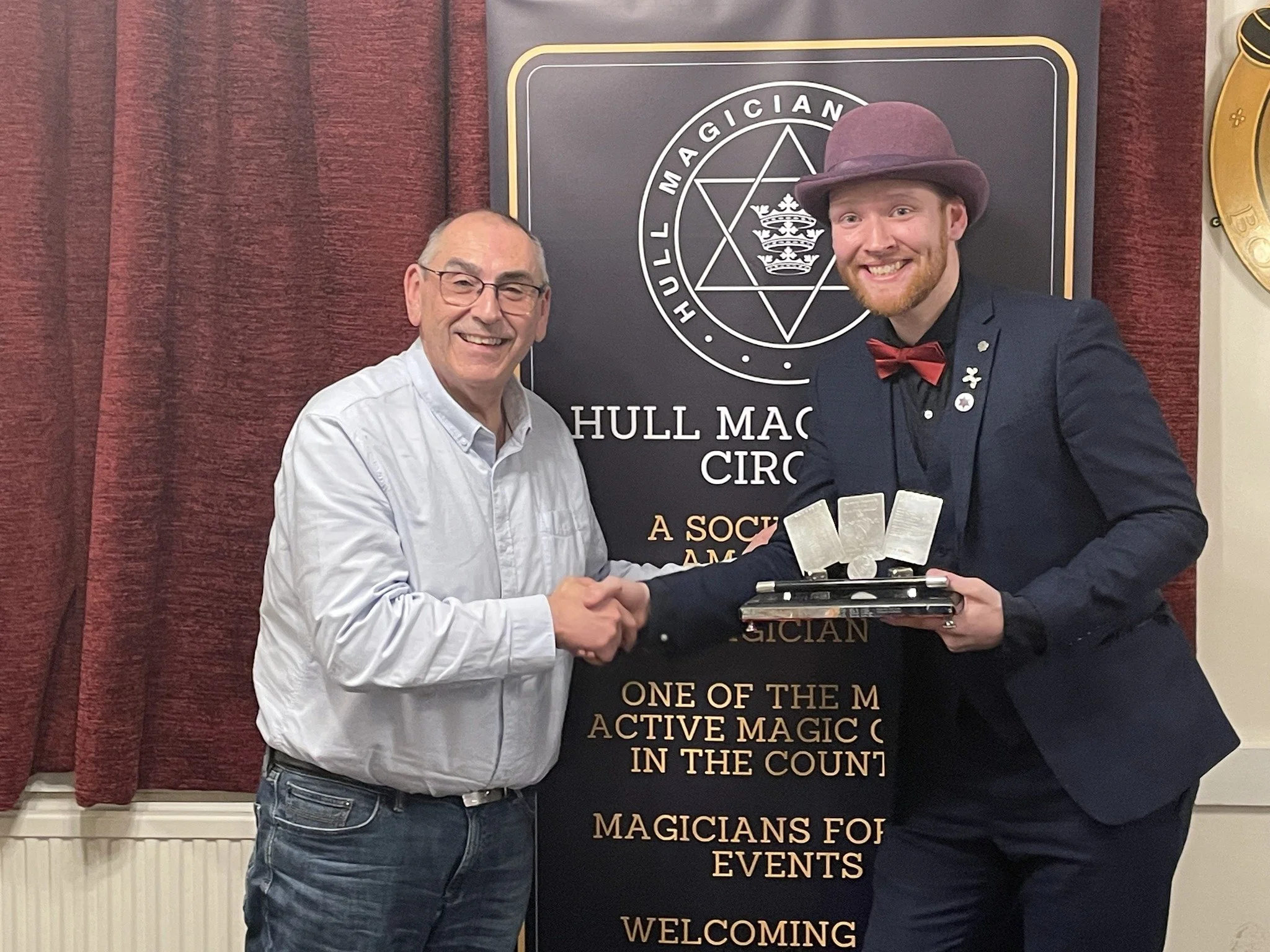 Two men shaking hands indoors, one of them wearing glasses and a white shirt, the other in a suit with a bow tie and a purple hat. They are smiling and holding a tray with four silver objects. Behind them is a banner with symbols and text related to magic and a red curtain.