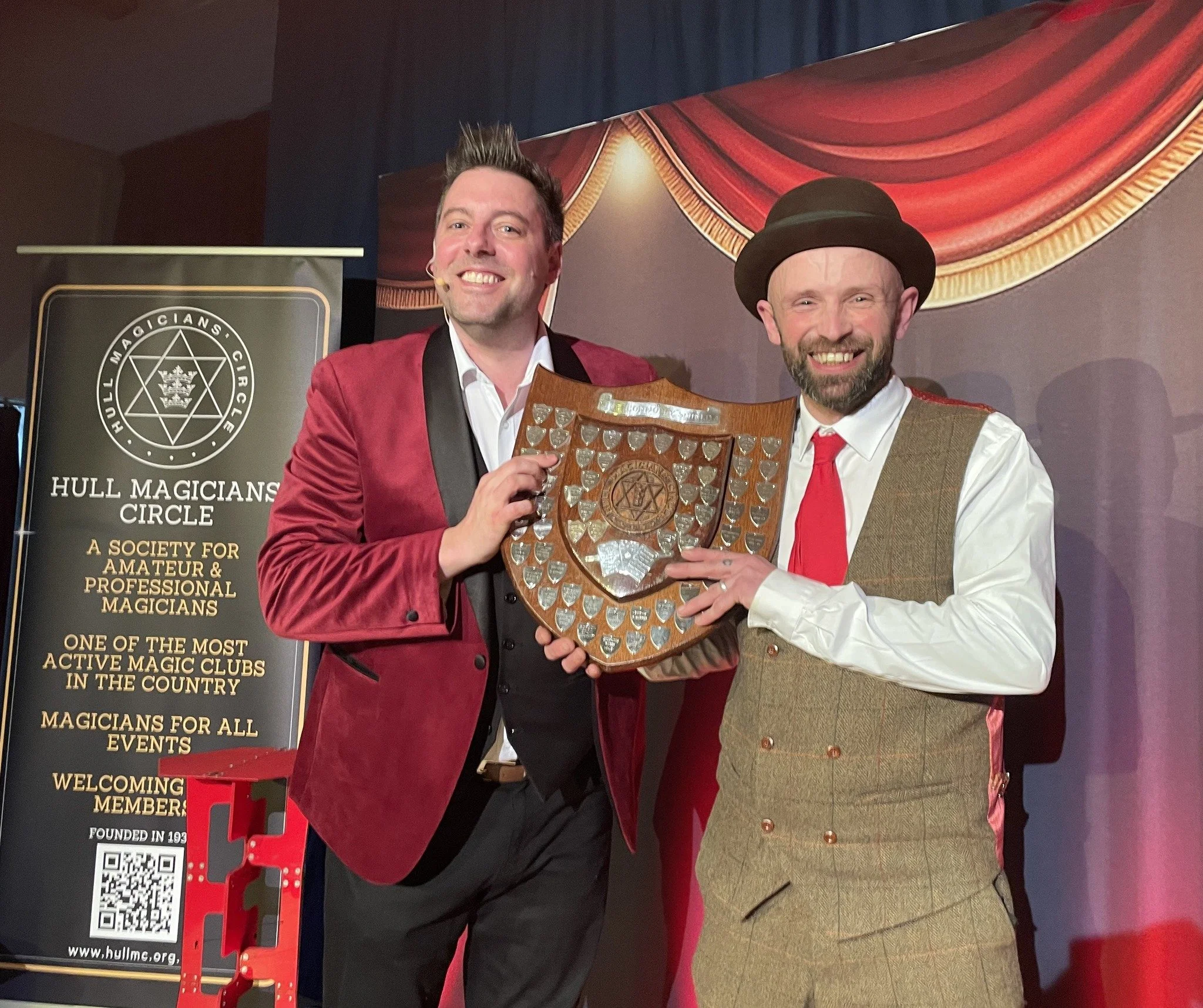 Two men in formal attire, one in a red velvet jacket and the other in vintage costume, holding a shield-shaped award with a star emblem and shield insignia, standing on stage in front of a banner that reads "Hull Magicians Circle."