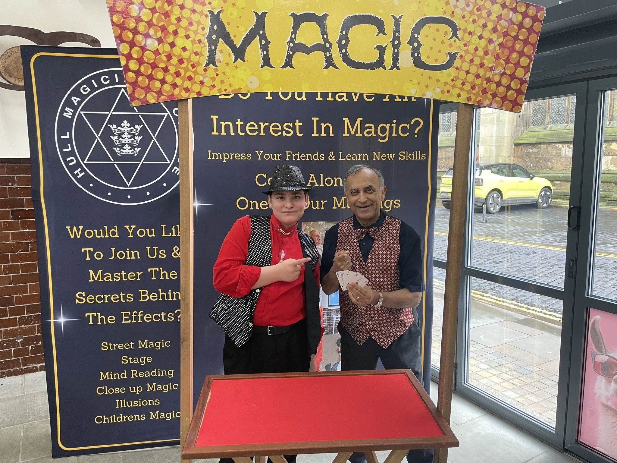 Two men in front of a magic show booth with a sign that reads "Magic". The younger man is wearing a red shirt, black vest, and hat, pointing at the older man. The older man is holding playing cards, smiling. The booth has a large blue banner with yellow text about magic tricks, and a smaller sign on top that says "Magic".