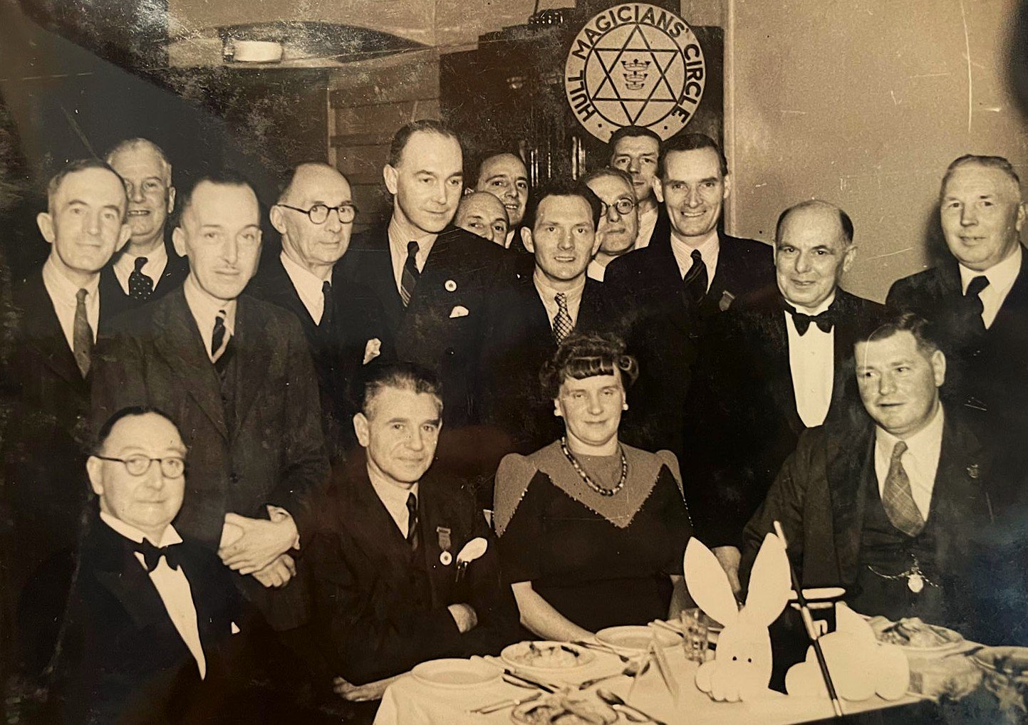 A group of men and women gathered at a dinner table at a social event, with a sign in the background reading 'Tulip Magicians' Circle,' suggesting a magician's club or society.