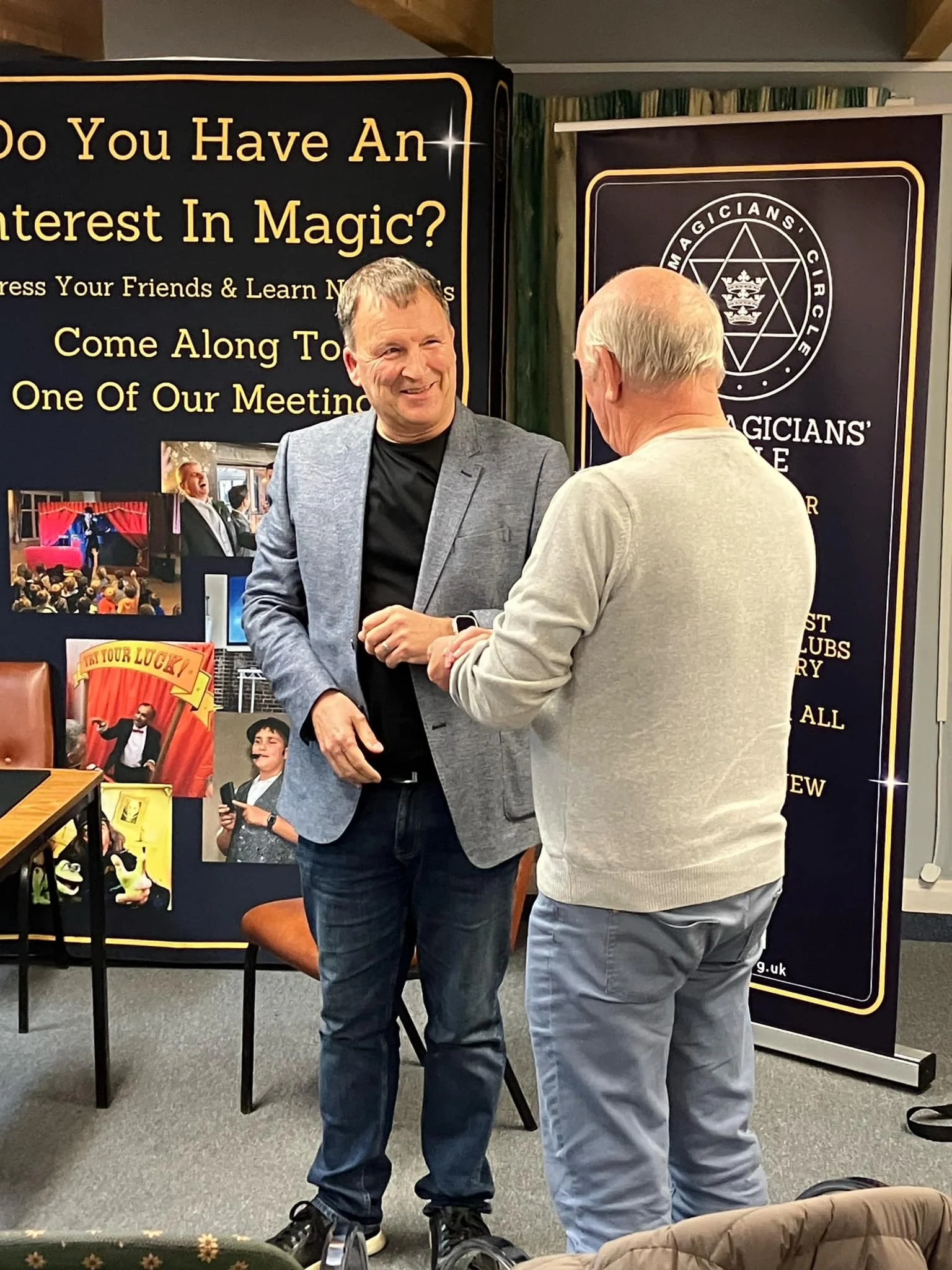 Two men shaking hands in front of a black and yellow promotional display for a magician's club, with photographs and text about magic community events.