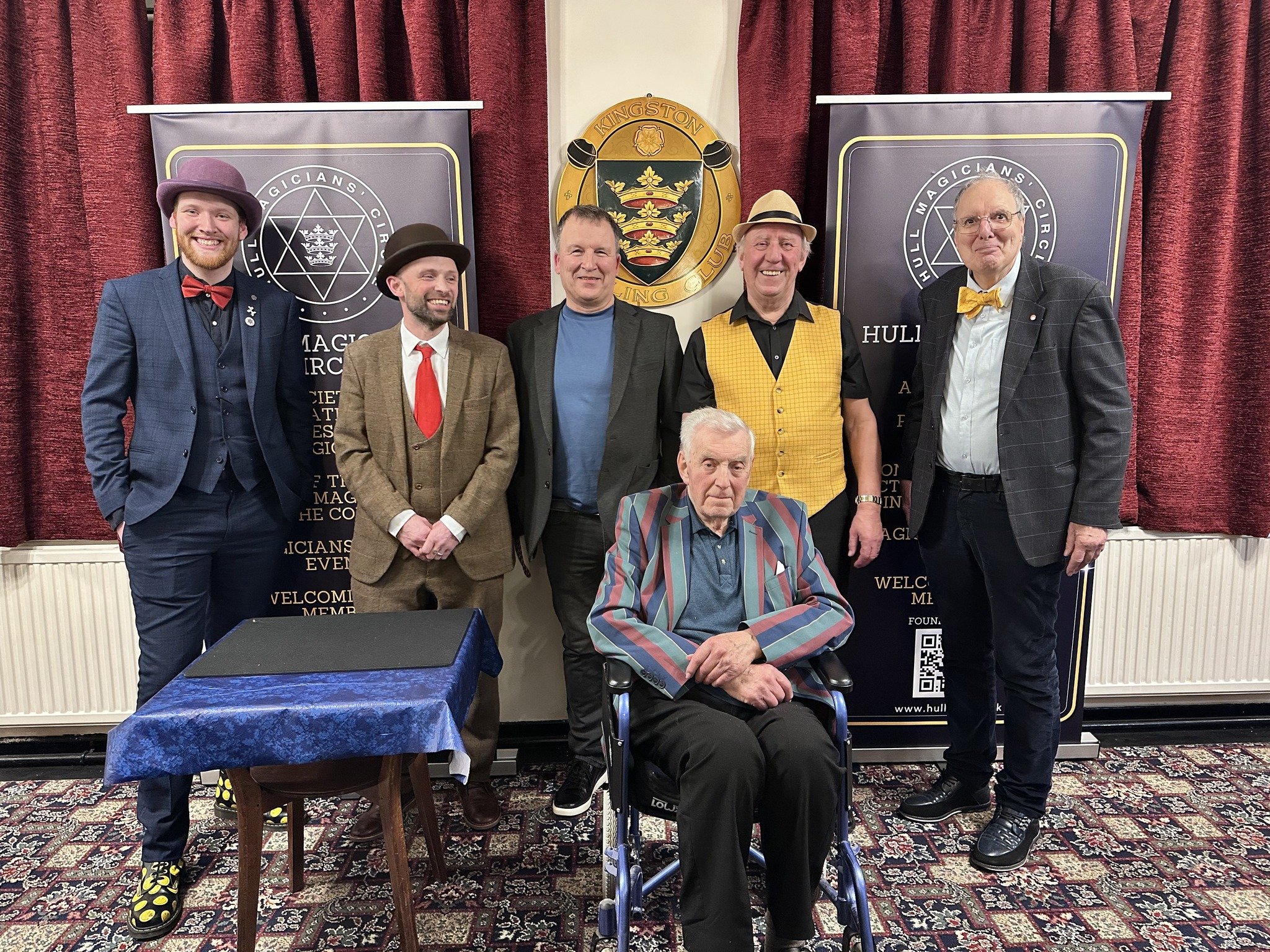 Group of six men, some with colorful bow ties and hats, standing behind an elderly man in a wheelchair in front of banners for the Hull Magicians Circus Club at an indoor event with red curtains.